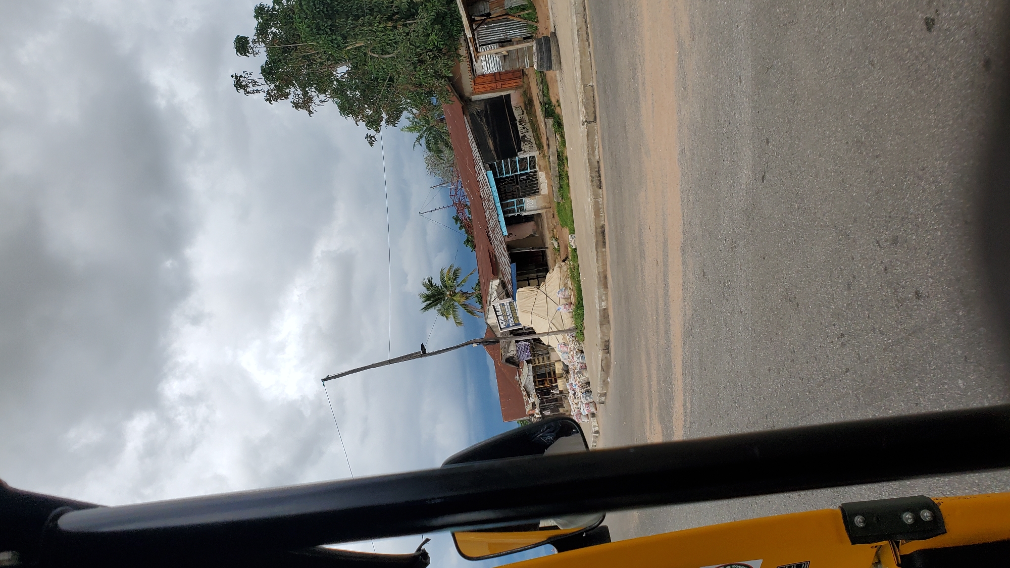The image, taken from a three-wheeled vehicle, shows a street scene in Jalingo, Nigeria, under a cloudy sky. In the foreground, the yellow body and black frame of the vehicle are visible. The road surface is asphalt, with some patches of sand and debris.
The middle ground features a row of shops with corrugated iron roofs, some appearing weathered. Bags of goods, possibly cement or produce, are stacked in front of some of these shops. A sign with text that appears to read "VICTOR GLAS" is prominently displayed. A palm tree stands behind the shops. A utility pole with wires leans precariously in front of the shops.
In the background, more trees and buildings are visible, along with a darker, stormier sky. The overall impression is of a market or commercial area on a potentially rainy day. There are no people clearly visible in the image.