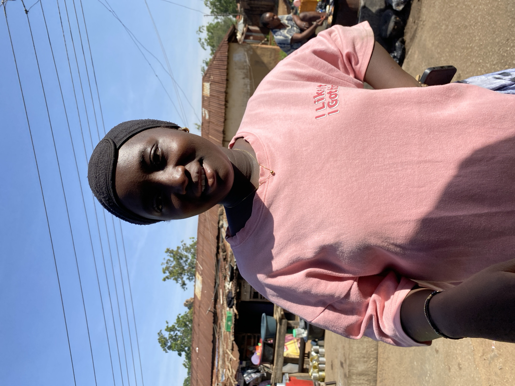 This image captures a young individual, likely female, posing outdoors under a clear blue sky in what appears to be Bukuru, Nigeria. The subject has dark skin and is wearing a black headband and a light pink long-sleeved shirt. She looks directly at the camera with a gentle smile, revealing some teeth. A thin gold necklace is visible around her neck, and a bracelet adorns her right wrist. In her right hand, mostly obscured, she holds a black smartphone.
The setting suggests a vibrant, semi-urban environment. In the background, structures with rusty corrugated metal roofs are visible, indicating homes or small businesses. Scattered in front of these buildings are various objects, including buckets, containers, and other items that might be market wares or household goods, suggesting an active area. A blurred figure is seated in the mid-ground, further hinting at ongoing daily activities.
The time of day is bright and sunny, with a clear blue sky devoid of clouds, indicating direct sunlight overhead. Several power lines crisscross the upper portion of the sky. The overall atmosphere appears casual and everyday.
Notable text on the subject's pink shirt reads "I Like It I Got It" in white letters.