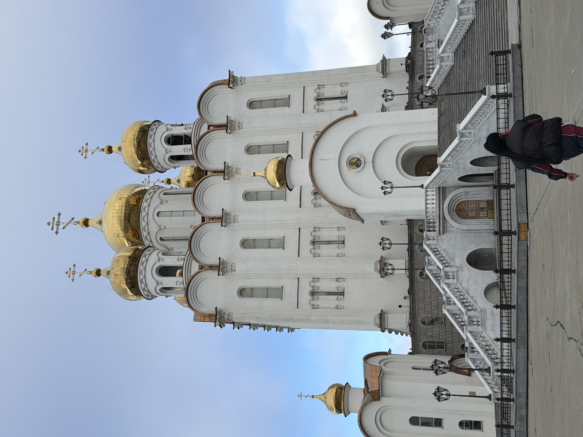 The image depicts a large, white, multi-domed Orthodox church with golden onion domes topped with crosses. The church has a grand white stone staircase leading up to an entrance. Several ornate street lamps are positioned along the staircase and the church grounds. In the foreground, a person wearing a dark jacket with red and blue stripes is walking away from the camera, down the paved area beside the stairs. The sky is a clear, pale blue with a few wispy clouds, suggesting it is daytime with fair weather. The architecture and the clear blue sky are consistent with a location in Magadan, Russia. No visible text is present in the image.