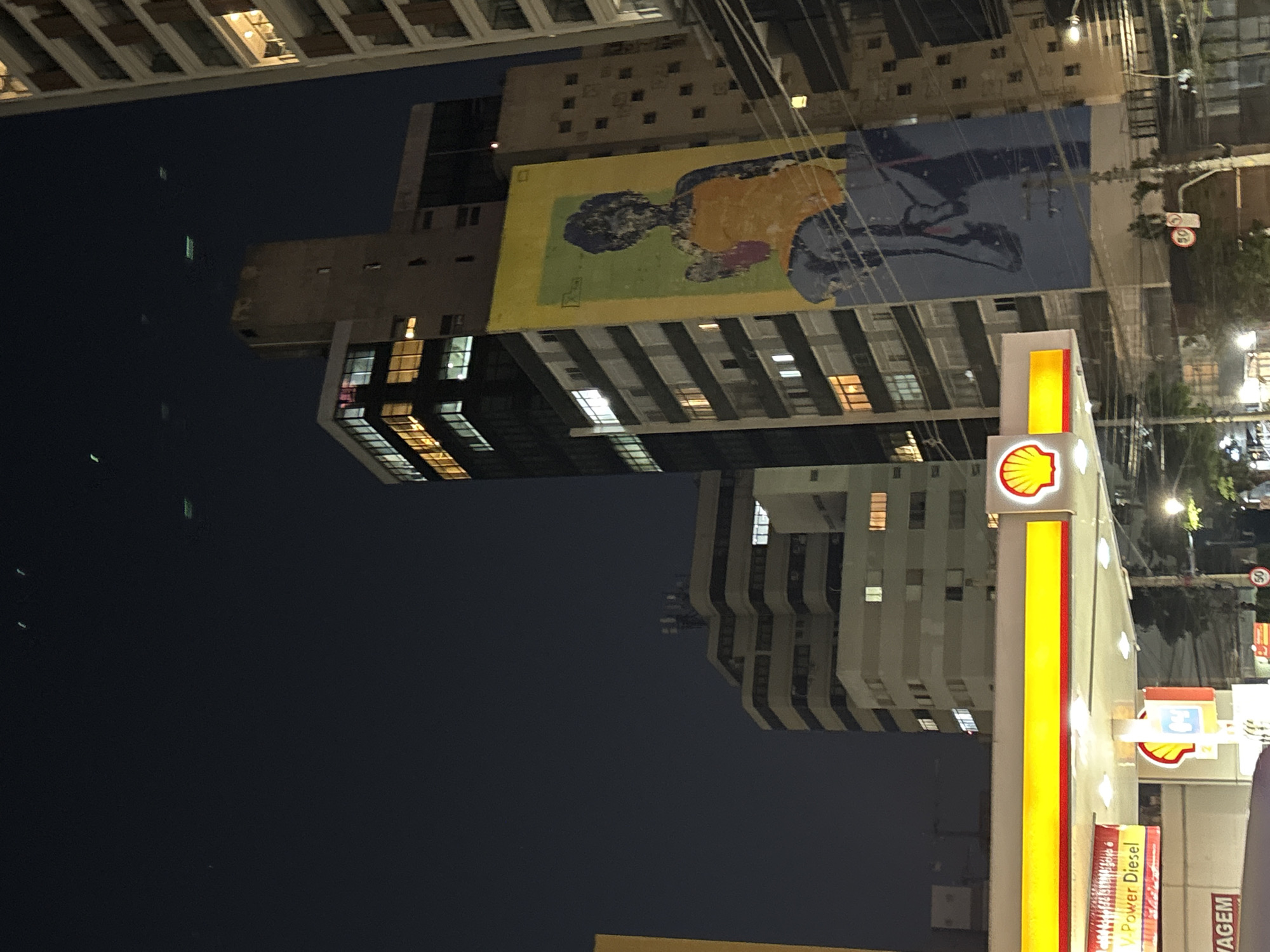 The image shows a nighttime view of a street scene in São Paulo, Brazil, likely near the Higienópolis neighborhood. In the foreground, a Shell gas station with illuminated signs is visible. The main sign prominently displays the Shell logo and reads "V-Power Diesel." Behind the gas station, several tall buildings rise into the dark sky. One building features a large mural of a woman in an orange dress and blue pants. Another building to the left has a series of illuminated windows, and a tall, slender building to the right also shows lit windows. The sky above is dark with several faint, blurry lights that could be distant aircraft or reflections. Traffic lights and streetlights are also present, casting a glow on the street. The overall impression is of a busy urban environment at night.