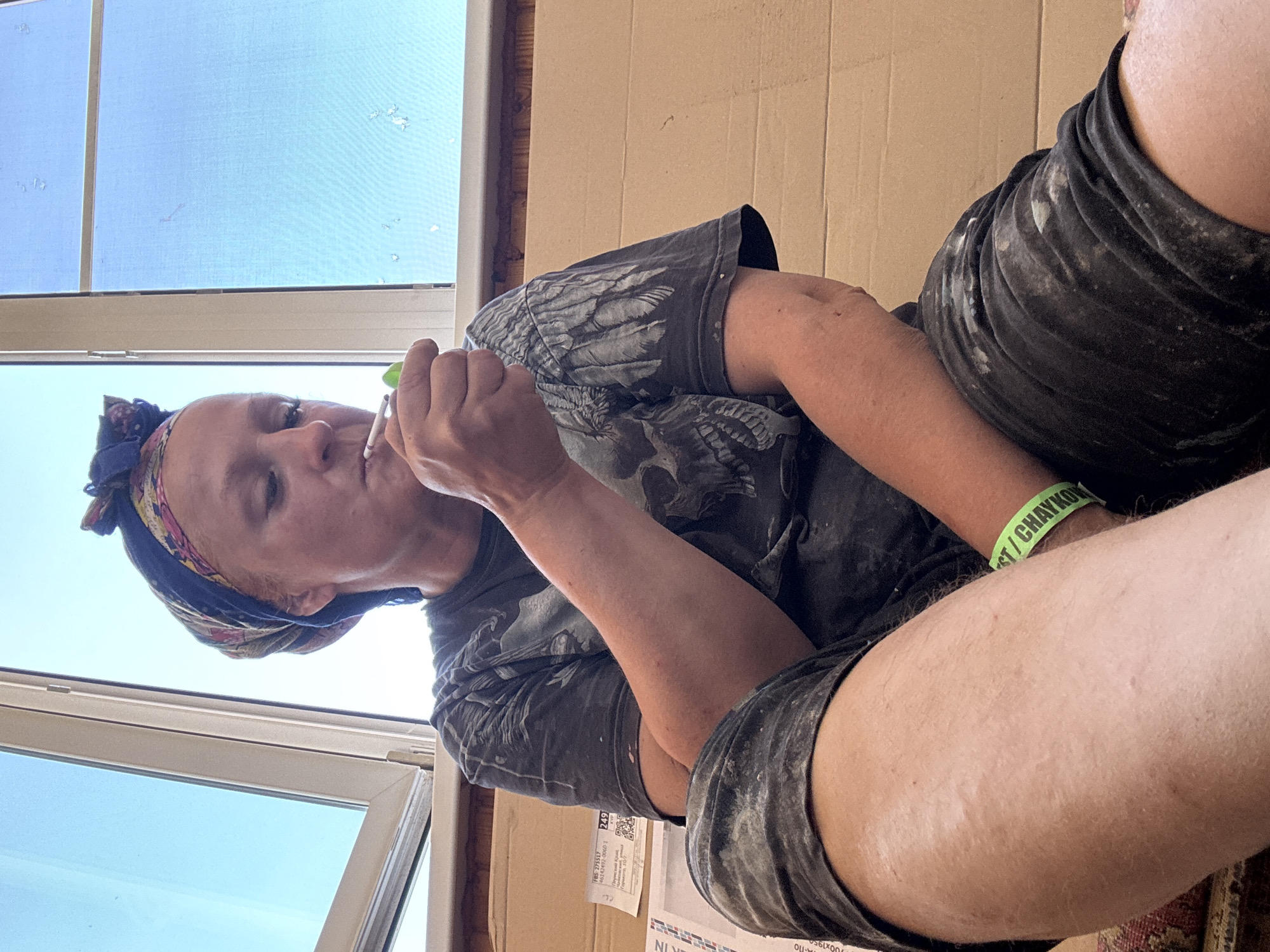 A seated individual wearing a headscarf, a t-shirt with a skull design, and shorts is holding a small, possibly lit, object to their mouth. The individual also wears a green wristband with writing that appears to include the word "CHAYKOVSKY," suggesting the location is the city of Chaikovsky in Russia. A window is visible to the left, revealing a clear sky. The person appears to be seated indoors, with a wall behind them.
