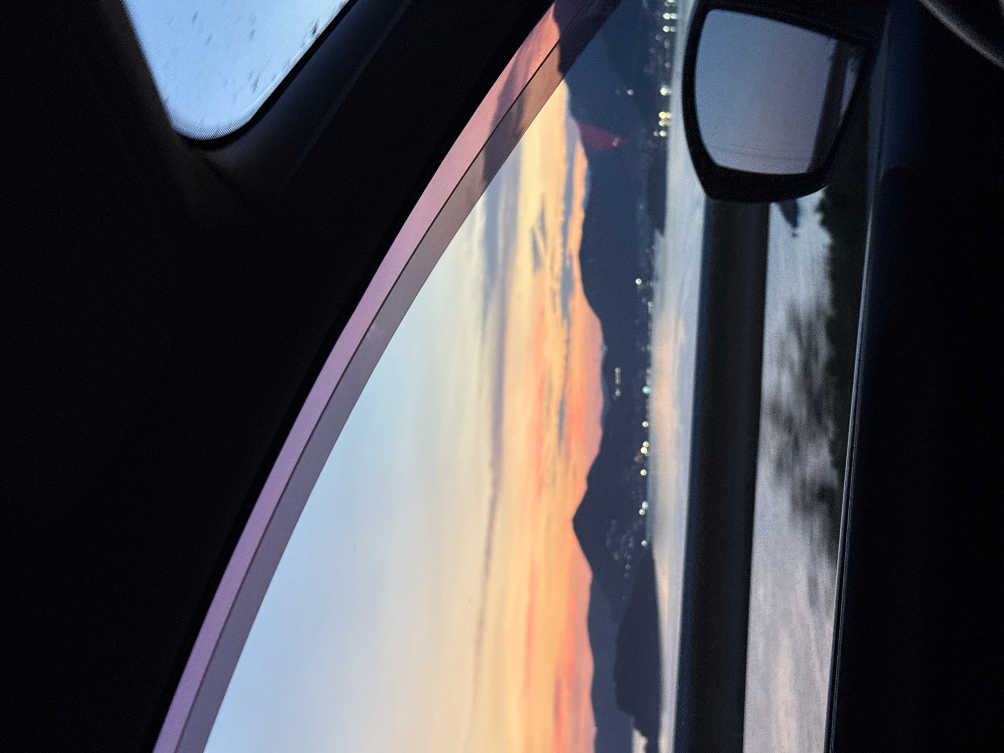 The image captures a scenic view from inside a vehicle at dusk. Through the car window, a body of water reflects the vibrant colors of the sunset, with hues of orange, pink, and purple painting the sky. Silhouetted mountains rise in the distance, their outlines softened by the fading light. Small, glowing lights are scattered along the coastline, suggesting a town or city nestled among the hills. A side-view mirror of the car is visible on the right, reflecting a portion of the serene twilight landscape. The interior of the car, including the window frame and part of the dashboard, frames the view, creating a sense of personal perspective.