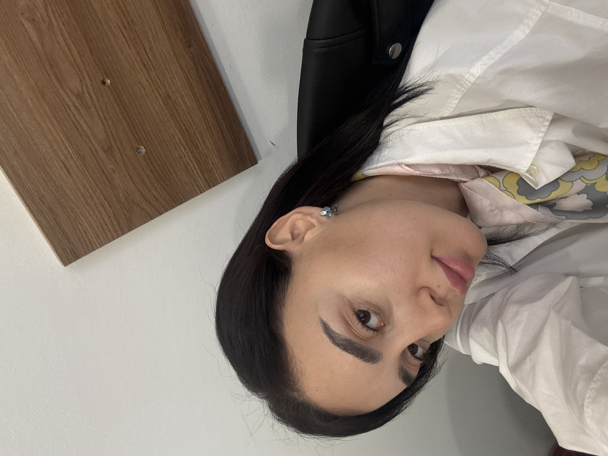 A medium shot shows a woman facing the camera with her head slightly tilted to the left. She has dark hair, and dark eyebrows, and is wearing a white shirt with a patterned scarf underneath. She also wears a blue and silver earring in her left ear. To her right is a black garment, possibly a jacket. To her left, there's a wooden panel attached to a white wall. The location and exact details of Tbilisi, Georgia are not evident in the image.