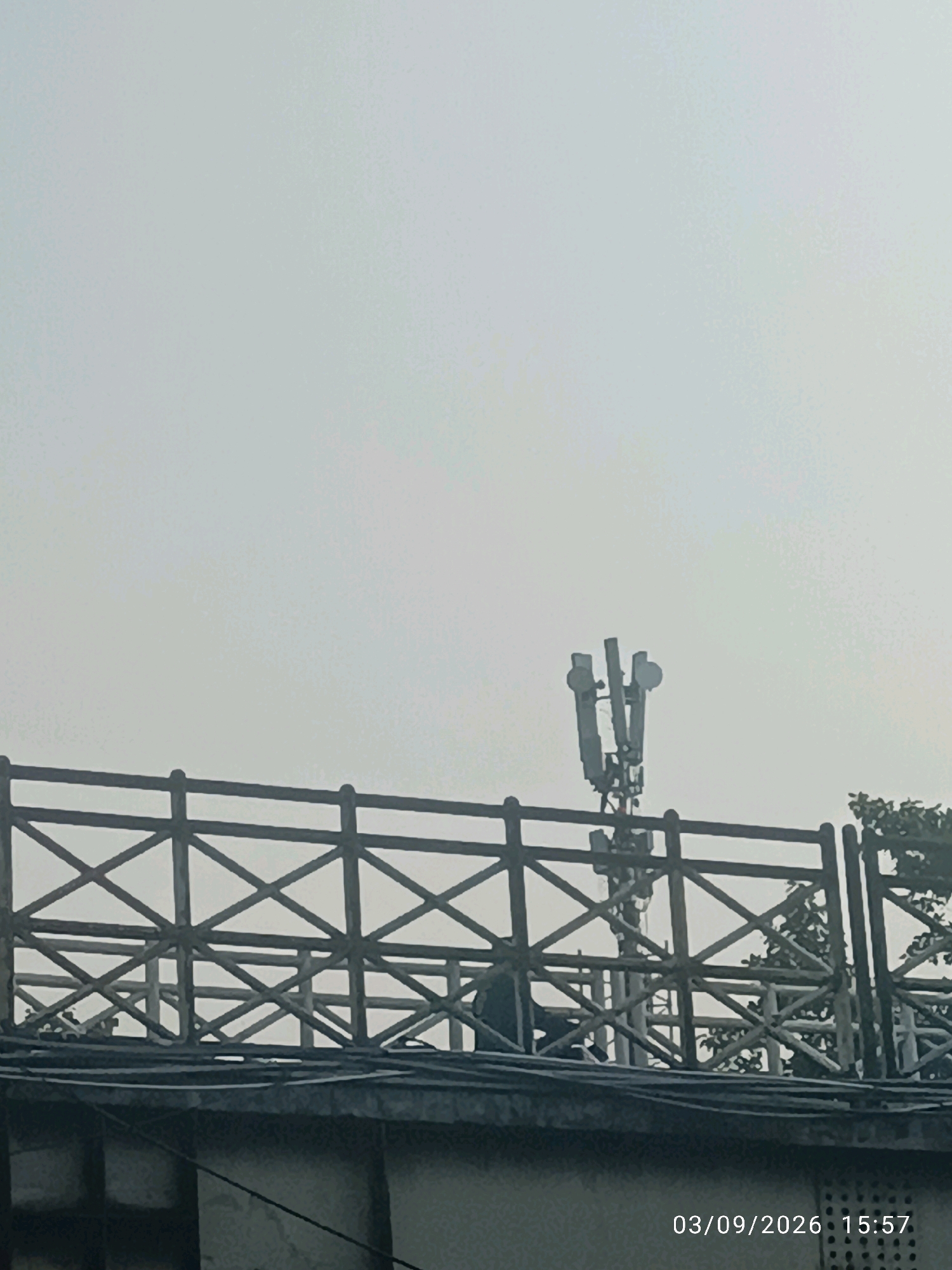 A cell tower stands against a hazy sky, positioned behind a wooden fence with a diamond-shaped lattice design. The fence is on top of a building, with some trees visible to the right. The lower part of the image shows a portion of a building with visible bricks and wires. A timestamp in the bottom right corner reads "03/09/2026 15:57".