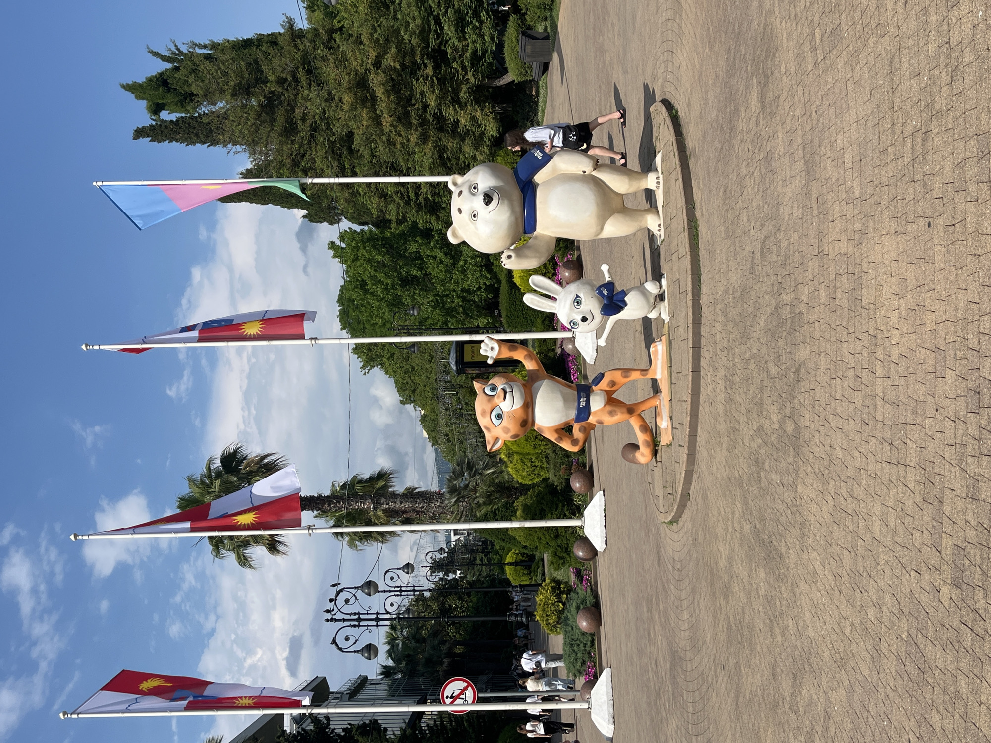 This media file showcases an outdoor scene in Sochi, Russia, likely during daylight hours given the bright sunlight and shadows. The setting appears to be a public walkway or park, paved with interlocking stones.
The primary subjects are large, colorful mascot statues of a polar bear, a rabbit, and a cheetah, all standing on a circular platform. The polar bear is wearing a blue scarf, the rabbit a blue bow, and the cheetah a blue band with the text "Sochi 2014" partially visible. These characters are likely related to the 2014 Sochi Winter Olympics.
In the background, several flags are displayed on poles. One flag has three horizontal stripes: blue, pink, and green. Two other flags are identical, featuring a red field with a white stripe and a yellow sun or star-like emblem. Lush green trees and palm trees are visible, suggesting a warm climate. A few people are casually walking in the background, one young woman in a white top and black shorts is particularly noticeable.
Streetlights with ornate designs are also present, along with some potted plants and benches. A "no stopping" sign with a stroller icon is visible near the bottom left, indicating specific regulations for the area. The overall mood seems relaxed and festive, consistent with a tourist or recreational area. The clear blue sky with scattered clouds suggests fair weather.