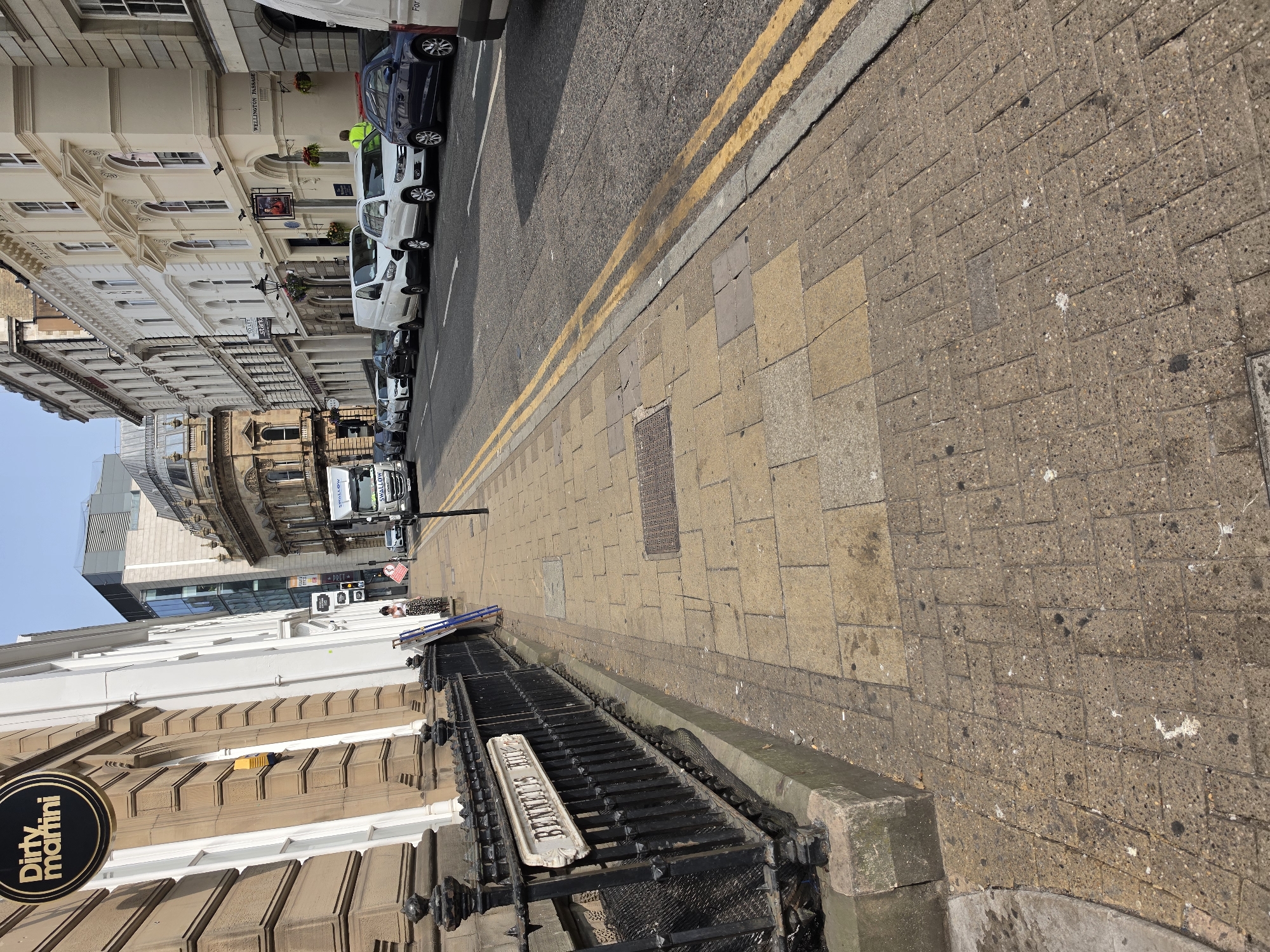The image captures a street scene in Birmingham, United Kingdom, with buildings lining the left side of the road and a sidewalk on the right. The perspective is from a slightly elevated position, looking down the street.
On the left, a row of buildings displays a mix of architectural styles. Some have ornate facades with classical detailing, large windows, and decorative cornices, painted in light cream and white. Further down, a more modern building with a glass facade is visible. A prominent sign with "Dirty Martini" in a circular logo is attached to one of the buildings.
The street itself has a tarmac surface with double yellow lines indicating parking restrictions. A line of parked vehicles, including white vans and dark-colored cars, is visible along the left side of the road.
The sidewalk on the right is paved with rectangular stone tiles. Several drain covers and a metal grate are embedded in the pavement. A black metal railing separates the sidewalk from a raised area, possibly a lower level or basement entrance, which has a white sign with ornate lettering that appears to read "BENNETT'S HILL".
A few people are visible in the image. One person in a yellow high-visibility vest is standing near the parked vehicles. Another person, wearing a patterned dress, is near a ladder that leans against a building. In the distance, a white truck with "SWALLON" written on it is driving away.
The environment appears to be a bright, sunny day with clear blue sky. The overall scene depicts a typical urban street with a combination of historic and modern architecture, commercial establishments, and vehicular traffic. The presence of the "Dirty Martini" sign and the street name "BENNETT'S HILL" strongly suggests the location is Bennett's Hill in Birmingham, a well-known area for bars and restaurants.