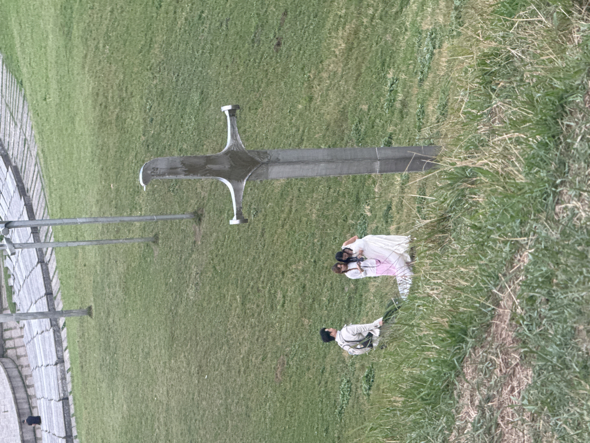 In Zemo Nichbisi, Georgia, a man wearing a light-colored jacket and a backpack stands on a grassy slope. He is looking to his right. To his left, two women are standing together. One woman is wearing a white dress and holding a camera, while the other woman is wearing a white top and a pink skirt. A large, metallic sword sculpture stands upright in the grassy area to the right of the group. In the background, a tiered stone seating area is visible, with a single person sitting on the seats. The environment appears to be an outdoor park or recreational area.