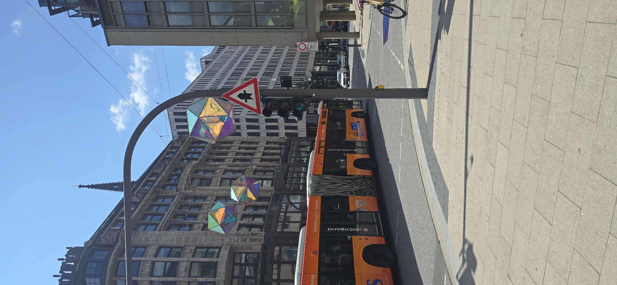 The image captures a street scene in Hamburg, Germany, specifically along the Mönckebergstraße, identifiable by the HOCHBAHN bus signage. A bright orange articulated bus, marked "HOCHBAHN" and "Deutschlands größte Fernschule," is prominent, traveling on the right side of the road. The bus is a modern model with multiple doors and advertisements on its side.
Suspended from an overhead curved metal structure are three large, geometric, iridescent decorations, casting colorful reflections. A traffic light pole stands to the right of the bus, featuring a red and white triangular warning sign indicating a merge or intersection ahead, along with the traffic signals. The sky is a clear blue with a few scattered clouds.
In the background, a mix of architectural styles is visible. To the left, a historic building with ornate stone facades and numerous windows stands, topped with a decorative cornice and a spire characteristic of the city's churches, likely St. Petri. To the right, a modern, multi-story building with a glass facade dominates the skyline. The street is paved with light-colored rectangular tiles, and a bicycle lane is delineated with gray and black textured tiles. A cyclist is partially visible on the right side of the frame, riding a bicycle. Cars are also present on the road in the background, suggesting urban traffic. The overall impression is a sunny day in a bustling European city.