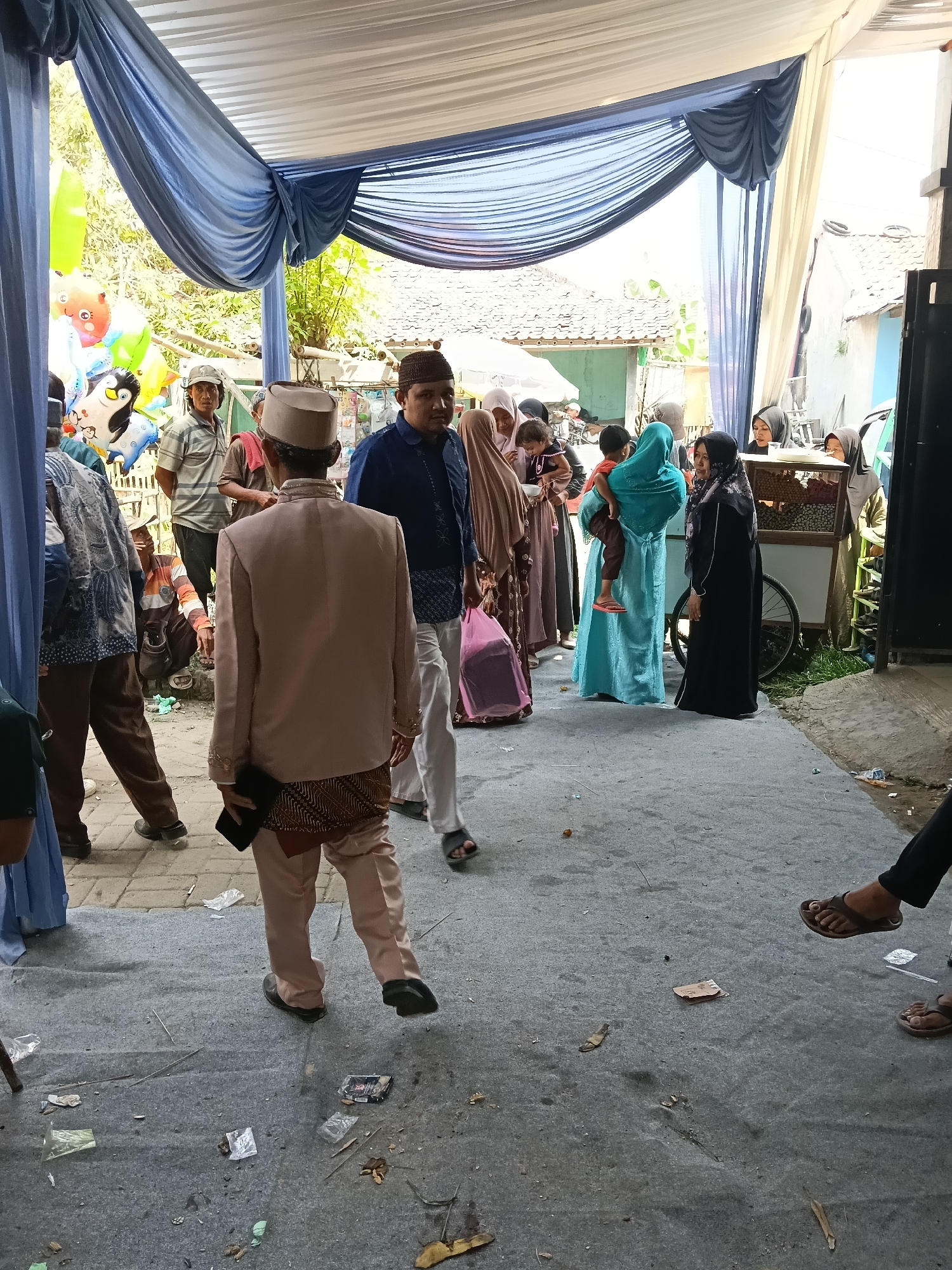 The image depicts an outdoor event, possibly a wedding reception, held under a temporary canopy with blue and white drapery. Several individuals are present, dressed in a mix of formal and casual attire. In the foreground, a man in a pale pink suit and a cap walks away from the camera, holding a black object. To his left, another man in a blue shirt and white pants is visible. To the left of the frame, people are gathered, some in traditional Islamic clothing, including women wearing hijabs. Balloons, including a character resembling Elmo and another that appears to be a penguin, are present on the left side, suggesting a celebratory occasion. In the background, there's a glimpse of a food cart with goods displayed and a glimpse of a building with a tiled roof. The ground is paved and shows some scattered debris, including plastic wrappers and leaves. The overall impression is a gathering of people in what appears to be a communal space in Tegal Angus, Indonesia.