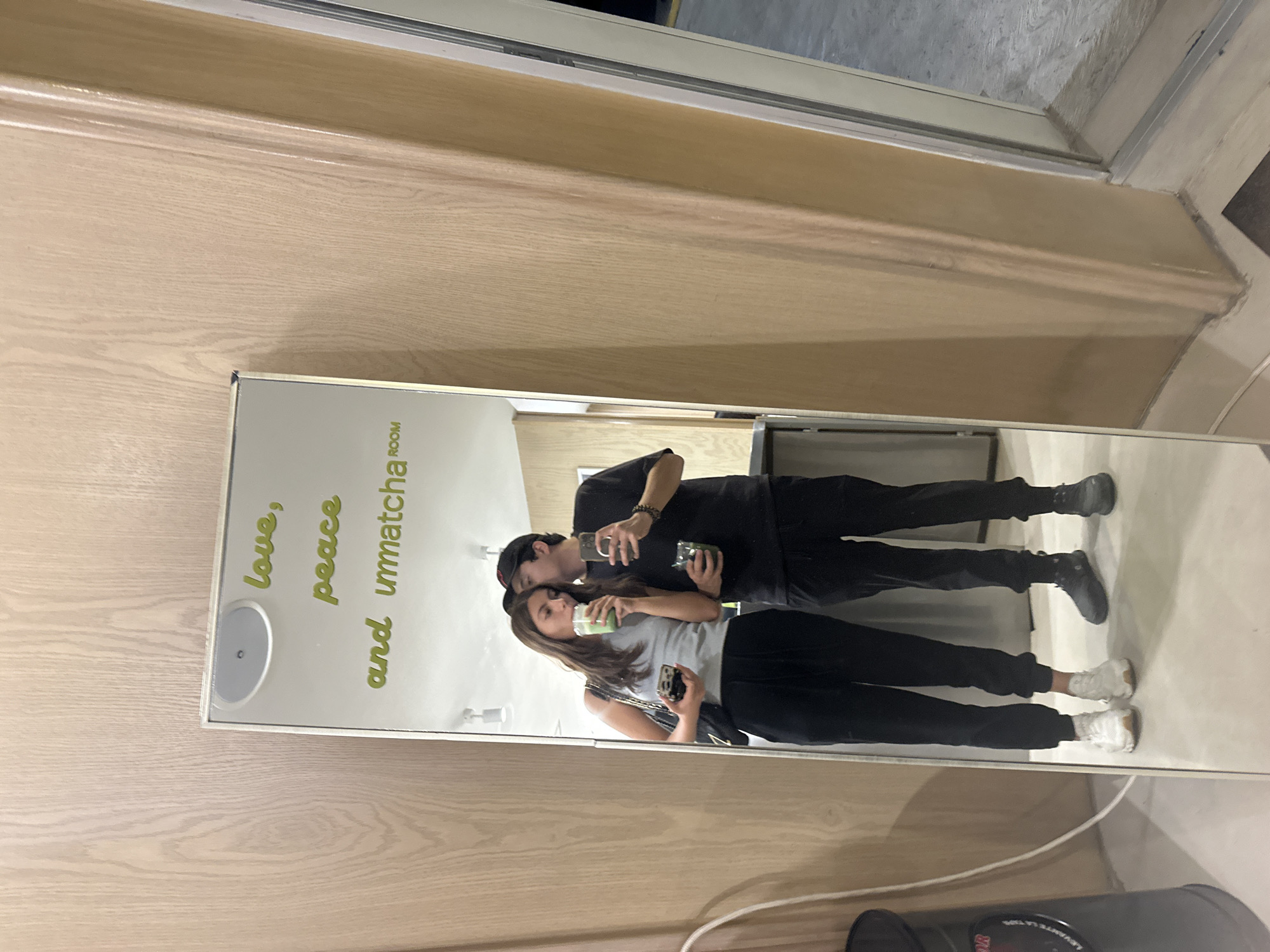 A couple is reflected in a mirror. The mirror is on a light wooden wall and has the words "love, peace and unmatcha room" printed on it in green. The couple is dressed in casual clothes, with the woman in a gray shirt and black pants, and the man in a black shirt and black pants. Both are holding drinks. They are standing close together, with the man's arm around the woman. The mirror is situated in what appears to be a modern space. The location is Puebla, Mexico.