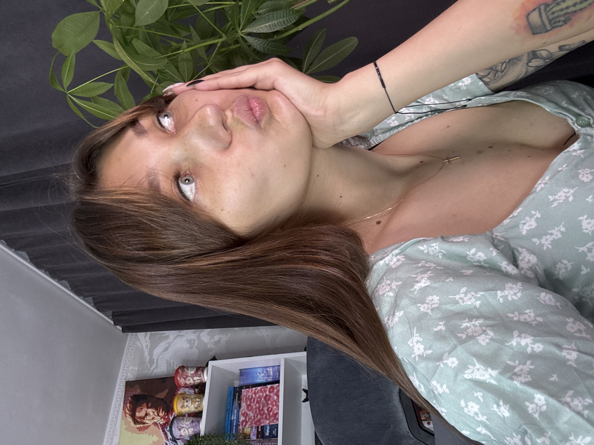 The image shows a young woman with long brown hair and freckles, looking to her right with her hand on her cheek and her lips pursed. She is wearing a light green dress with a white floral pattern and a gold cross necklace. A black beaded bracelet is visible on her wrist, along with tattoos on her arm. In the background, a bookshelf filled with books and decorative items is partially visible, including a poster of Tony Stark. A large green plant is to the right of the woman. The scene appears to be indoors, possibly in a home or office setting. There is no discernible information in the image to indicate the location in Novokuznetsk, Russia.