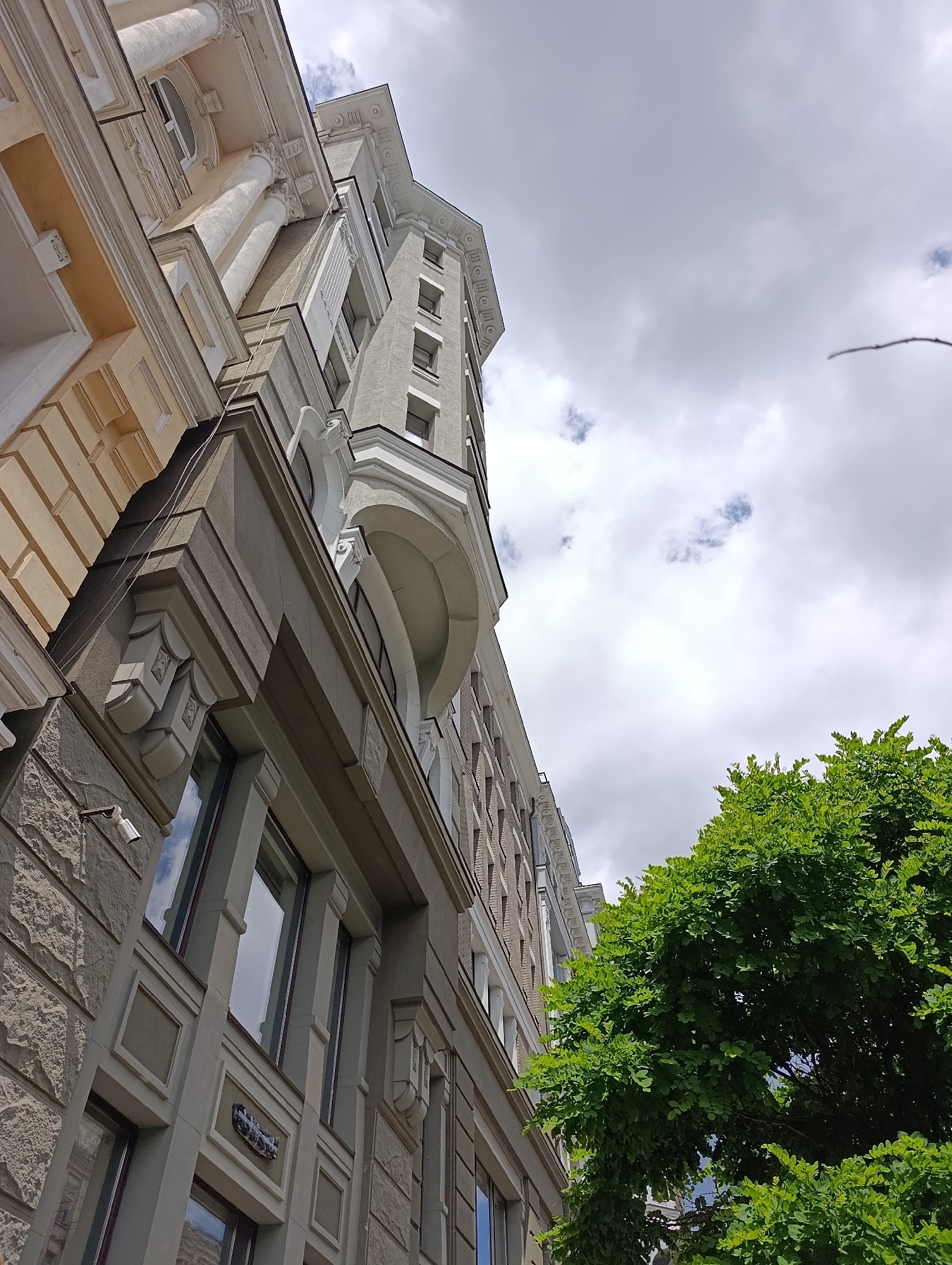 The image shows an upward-angle view of an old building in Kyiv, Ukraine, with a cloudy sky in the background. The building exhibits classical architectural elements such as columns, ornate moldings, and detailed facade work. The facade is a mix of light yellow and gray stone or stucco. A security camera is mounted on the stone facade on the lower left. To the right, lush green trees partially obscure the upper part of the building. The scene suggests a daytime setting in an urban environment.