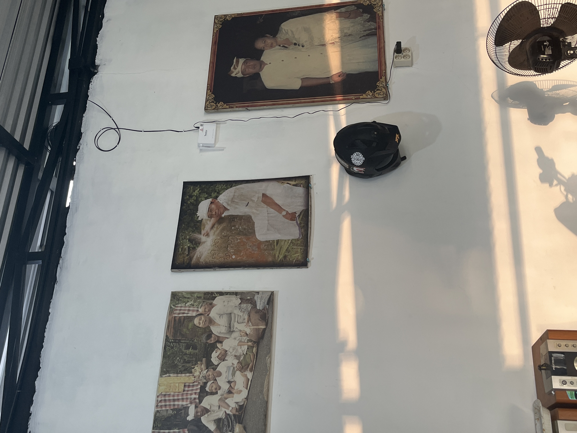 The image captures a section of a white wall adorned with framed photographs and various objects, suggesting an indoor or semi-outdoor space. On the left, structural elements with black metal beams and corrugated material are visible, contrasting with the smooth white wall.
Three framed pictures are mounted vertically on the wall. The topmost features a formal portrait of an older couple dressed in light-colored traditional attire. The middle photo depicts a person, also in traditional white clothing and headwear, seemingly engaged in a craft or ritual involving a stone object. The bottom frame displays a group of people, possibly a family or community, seated on a mat, also in traditional Balinese-style clothing, smiling towards the camera.
Several other items are attached to or placed near the wall. A white rectangular electronic device, possibly a modem or router, is fixed to the wall with black electrical wires extending from it, one coiled neatly. A black bicycle helmet with "JPX" visible on it hangs below the top photo. To the right, an electrical outlet with two sockets is mounted, and a dark wall-mounted fan is visible in the upper right corner.
Strong, elongated shadows are cast across the white wall from the right side, indicating direct sunlight, likely from a window or opening, suggesting late afternoon or early morning. The fan also casts a distinct shadow. In the bottom right corner, part of a wooden stand holds an electronic appliance with "POLYTRON" clearly visible, which appears to be a sound system or coffee maker.
The traditional attire seen in the photographs and the general setting align with the cultural context of Tegallalang, Indonesia, specifically Bali.
