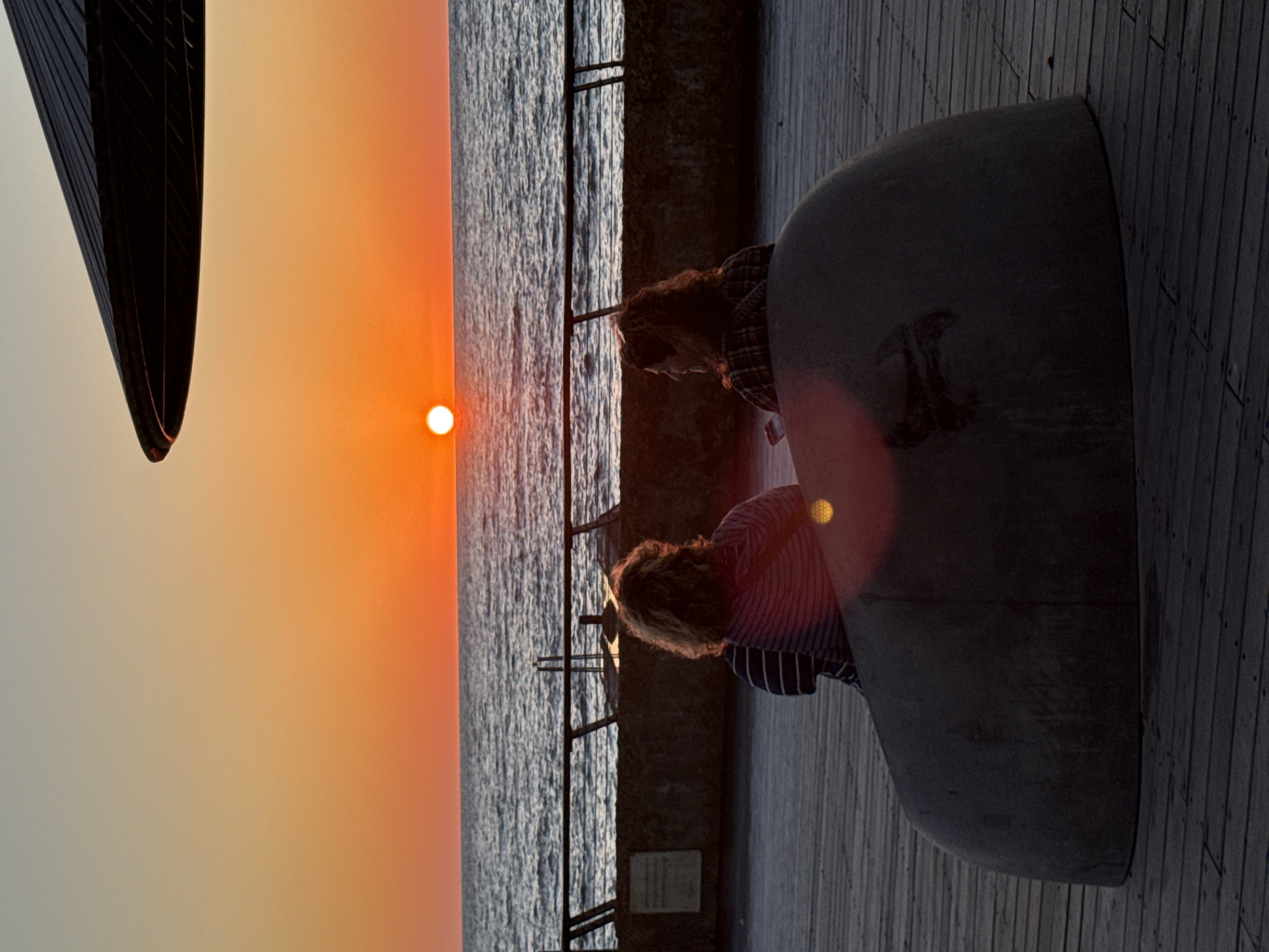 This photograph captures a tranquil sunset scene along a promenade in Tel-Aviv, Israel, with the image rotated 90 degrees counter-clockwise, placing the horizon vertically.
The primary subjects are two individuals, seen from behind and slightly to the side, seated on a dark, curved, bench-like structure. The person closer to the foreground has lighter, possibly blonde or light brown, curly hair and is wearing a blue and white striped shirt. The person seated behind them has reddish-brown curly hair and is wearing a plaid shirt. They appear to be observing the setting sun and the sea.
The activity taking place is quiet contemplation or relaxation, as the two people watch the sunset. The time of day is clearly evening, marked by the sun, a distinct bright white sphere, low on the horizon. The sky gradient transitions from a vibrant orange and yellow hue near the sun to a lighter, almost white-grey tone further away.
To the left of the seated individuals is the expanse of the sea, its surface textured with small waves, bordered by a dark concrete wall or railing. On the right side of the frame, a wooden deck or walkway is visible, indicating a promenade setting. In the far upper left corner, a large, dark, architectural structure, possibly a building or monument, is silhouetted against the bright sky. A subtle lens flare emanates from the sun's position. On the dark bench, near the person in the plaid shirt, there is a faint, dark marking or symbol. A light-colored rectangular plaque or sign is affixed to the wall behind the people, but any text on it is indistinct. The overall atmosphere is calm and contemplative.