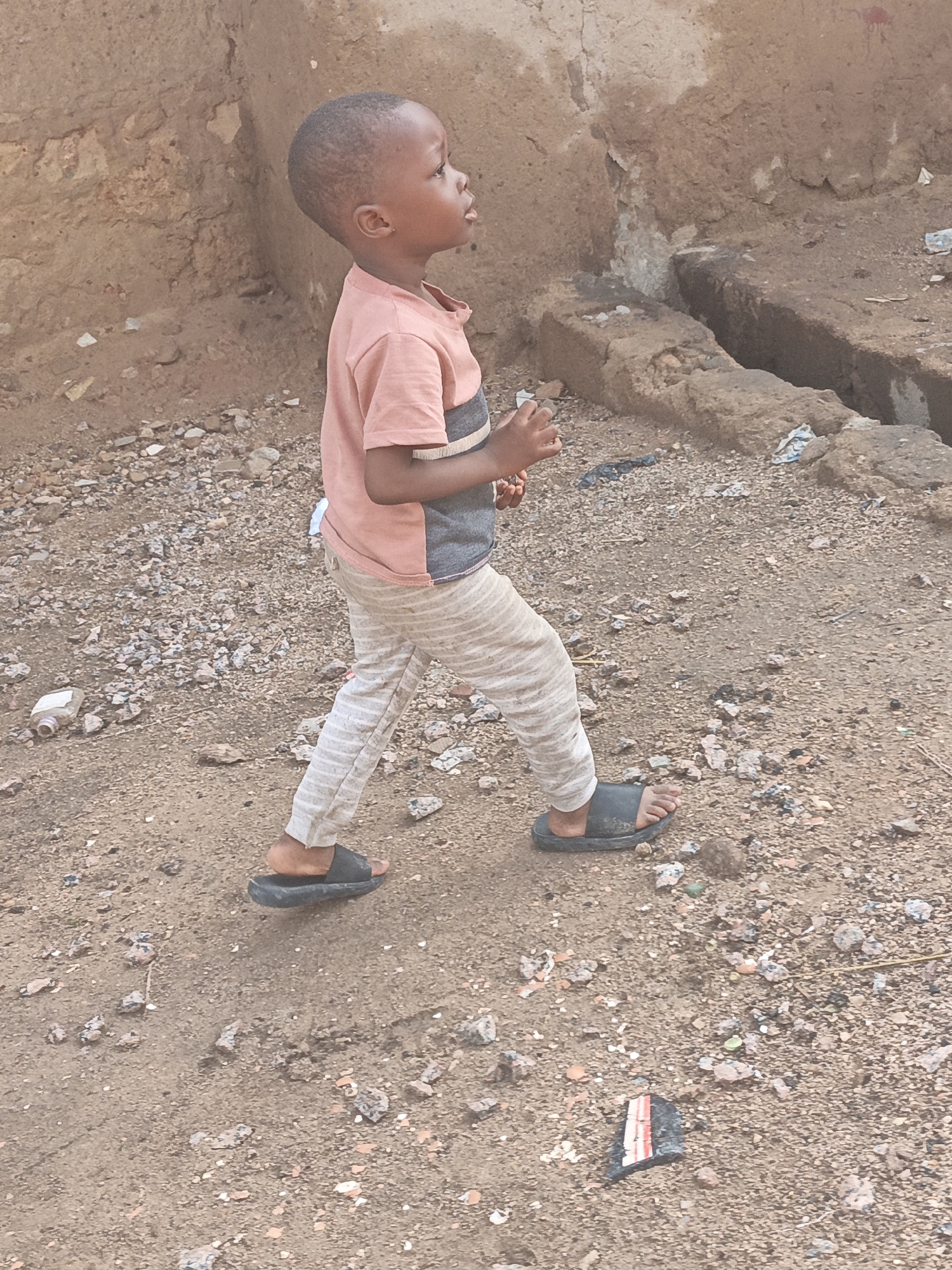 A young boy, appearing to be of African descent and wearing a pink and grey t-shirt and striped pants, walks on a dirt and gravel path in Gwagwarwa, Nigeria. The boy is facing to the right, looking upwards. His left foot is shod in a black sandal, while his right foot is bare and is also in a black sandal. The background consists of a rough, textured earthen wall and a drain or channel carved into the ground. Scattered on the ground are small rocks, pebbles, and some debris, including a discarded plastic bottle and fragments of what appears to be paper or plastic. The overall environment suggests an outdoor setting in a potentially unpaved or developing urban area.