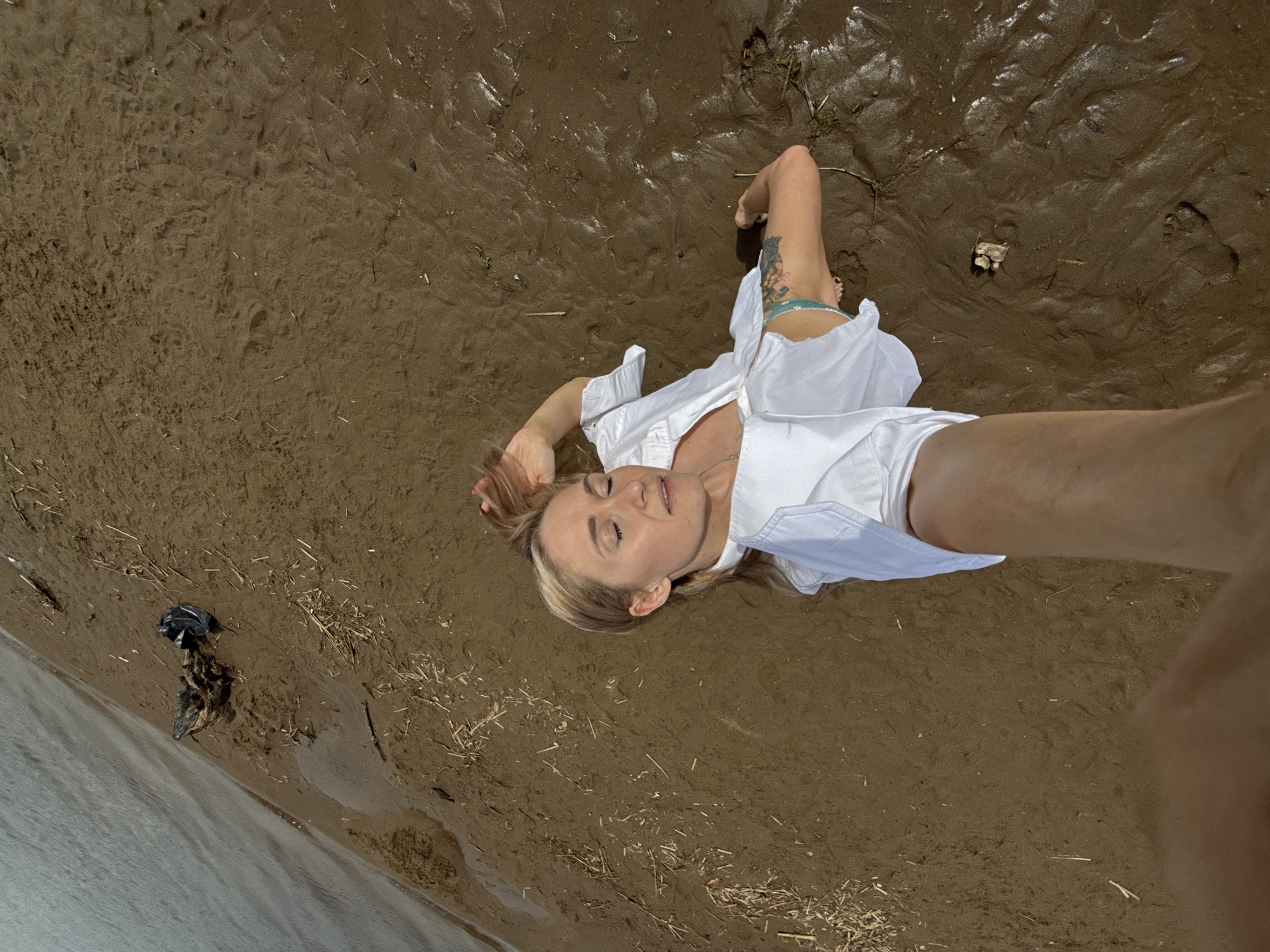 A fair-skinned, light-haired woman lies on a sandy beach in Chaikovsky, Russia. She wears a white, unbuttoned shirt and light-colored shorts with a tattooed leg exposed. She is lying near a body of water, and the ground is a mixture of wet and dry sand, with some debris such as twigs and a black plastic bag nearby. The picture is a selfie, taken from above, with the person's arm visible in the frame.