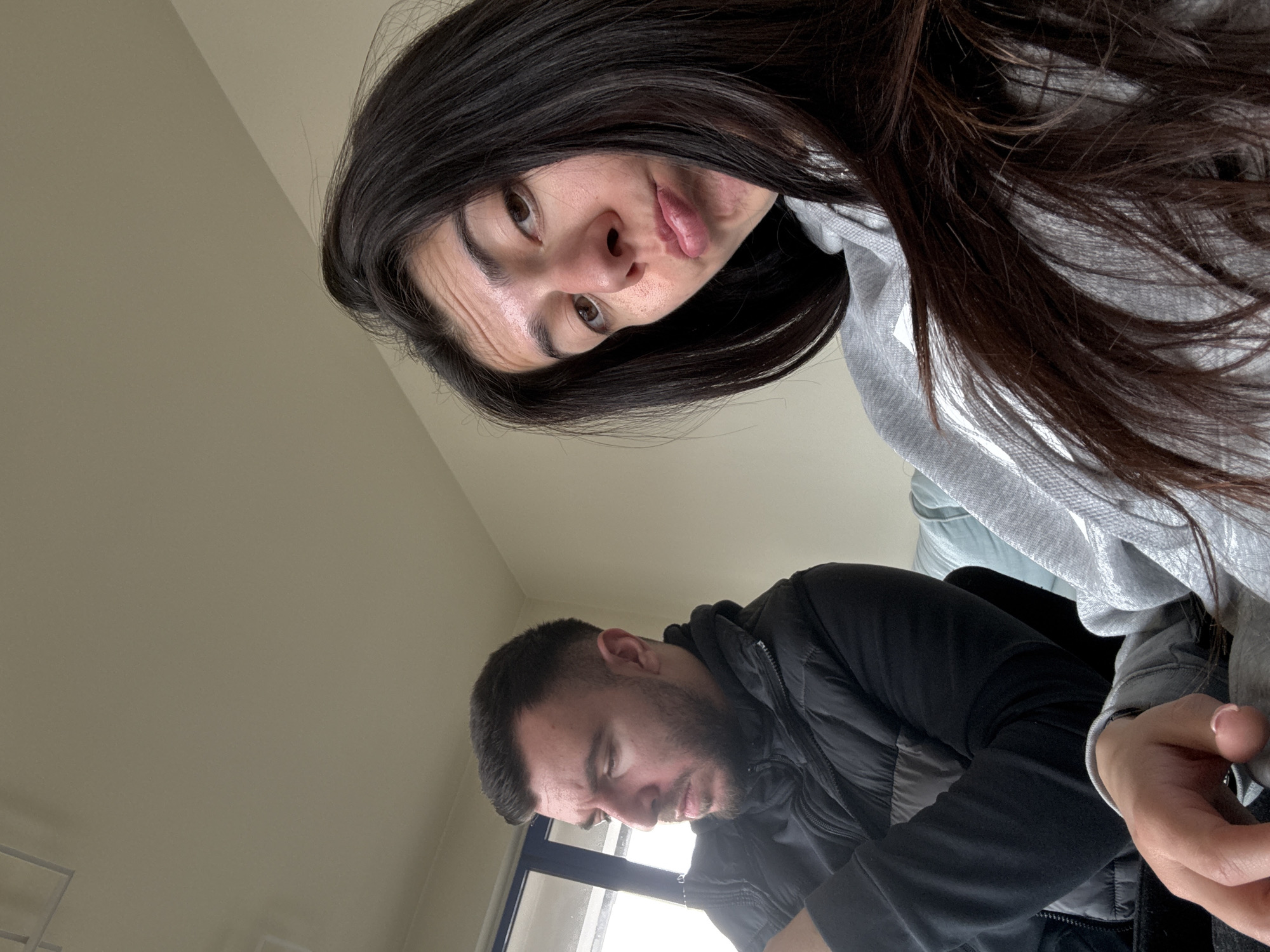 The image depicts two individuals in what appears to be an indoor setting. A woman with long, dark hair is positioned in the foreground, looking down at the camera. She is wearing a light-colored hooded sweatshirt. Below her, a man with short hair and a beard is looking down, possibly at a table or desk. He is wearing a black jacket. The background shows a light-colored wall and a window. The location in the city of Sofia, Bulgaria cannot be determined from the image.