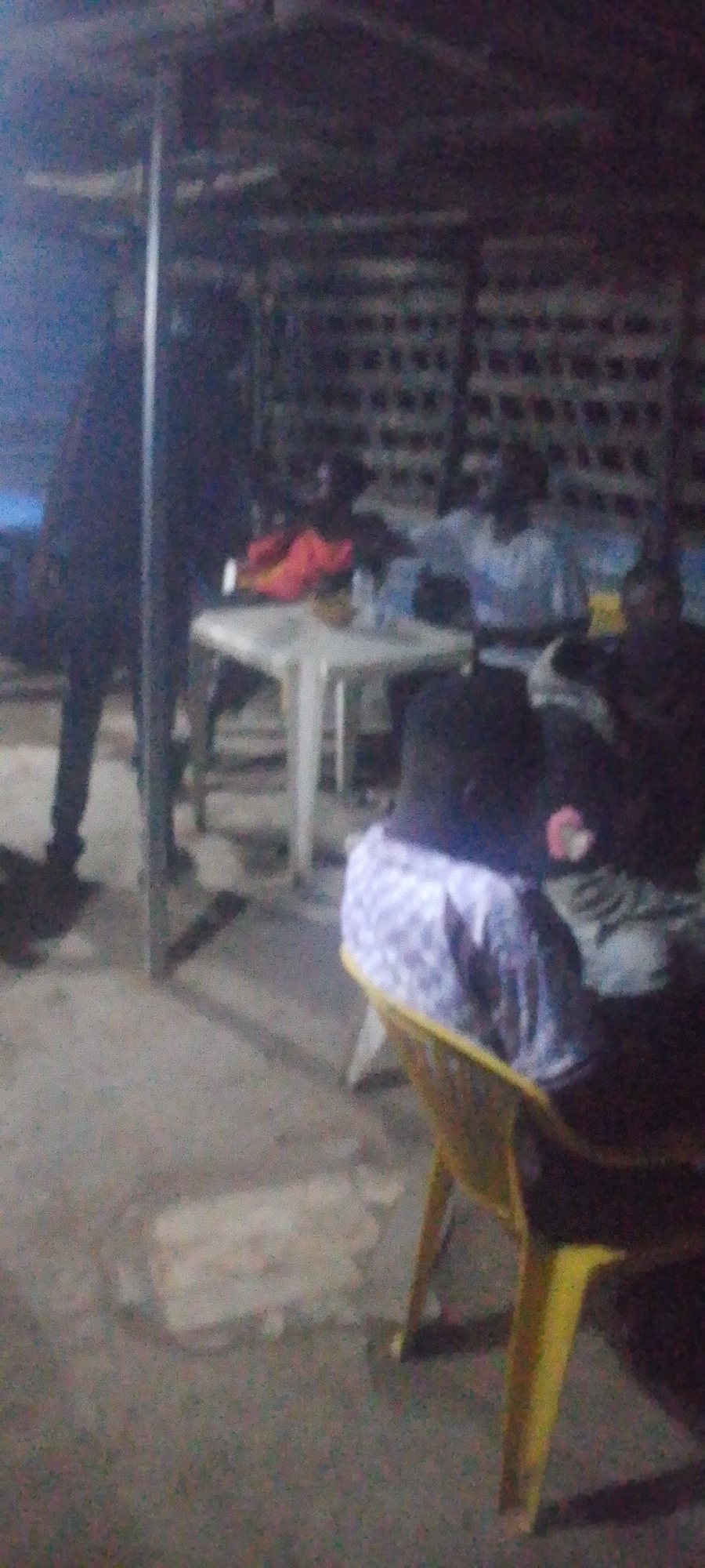 A group of people are gathered around a white table in what appears to be an outdoor or semi-outdoor seating area. The individuals are seated on chairs, some of which are yellow plastic. On the table are several bottles of water and what looks like food. The background is dimly lit with what appears to be brick or block walls. The overall scene suggests a casual gathering or meal.