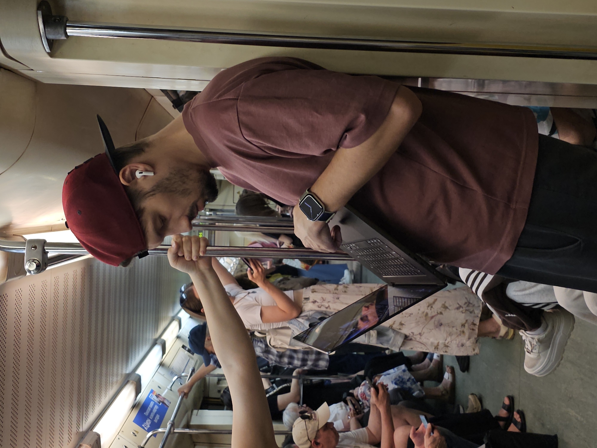 In a subway car, a man wearing a burgundy t-shirt, dark pants, a burgundy baseball cap, and white earbuds stands with his back to a pole, using a laptop that is angled upwards. He wears a smartwatch on his left wrist. In the background, other passengers are seated and standing. A woman in a white top and floral skirt holds her phone, and another person with a blue cap and plaid shirt is visible. The subway car interior features white ceilings with perforated panels, overhead fluorescent lighting, and silver poles.