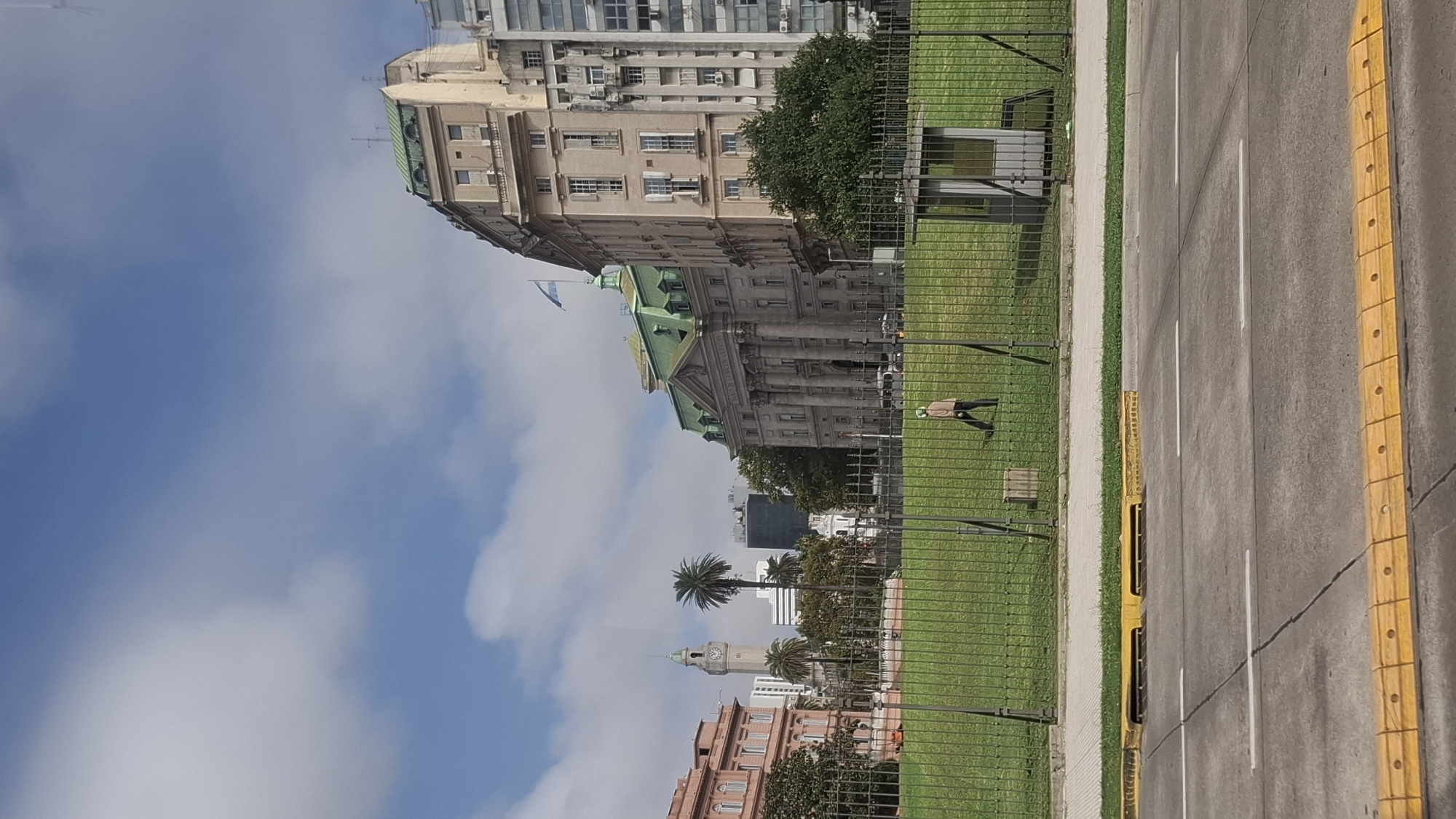 The image shows a street in Buenos Aires, Argentina. In the background are several buildings, including the Presidential Palace, known as the Casa Rosada (Pink House), identifiable by its pink facade and clock tower. In the midground is a green lawn, fenced off from the street, where a lone person is walking. The street itself has white lane markings and yellow speed bumps. A clear blue sky with scattered clouds provides the backdrop.