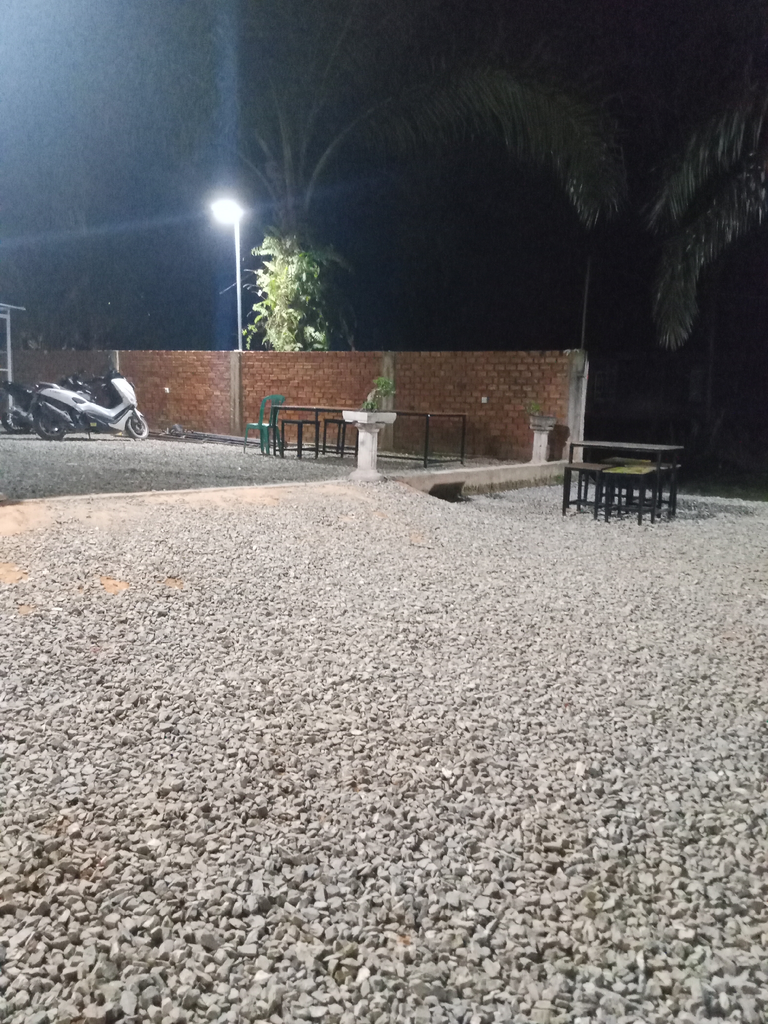 The image depicts an outdoor seating area at night in Mekar Jaya, Indonesia. A gravel ground covers the foreground and extends towards a brick wall in the background. A street lamp illuminates the scene. To the left, several motorcycles are parked. In the middle ground, a green chair is positioned next to a set of dark tables and chairs. A stone structure, possibly a fountain or pedestal, is located to the right of the tables and chairs. Palm trees are visible against the dark night sky. The area appears to be a casual dining or gathering space.