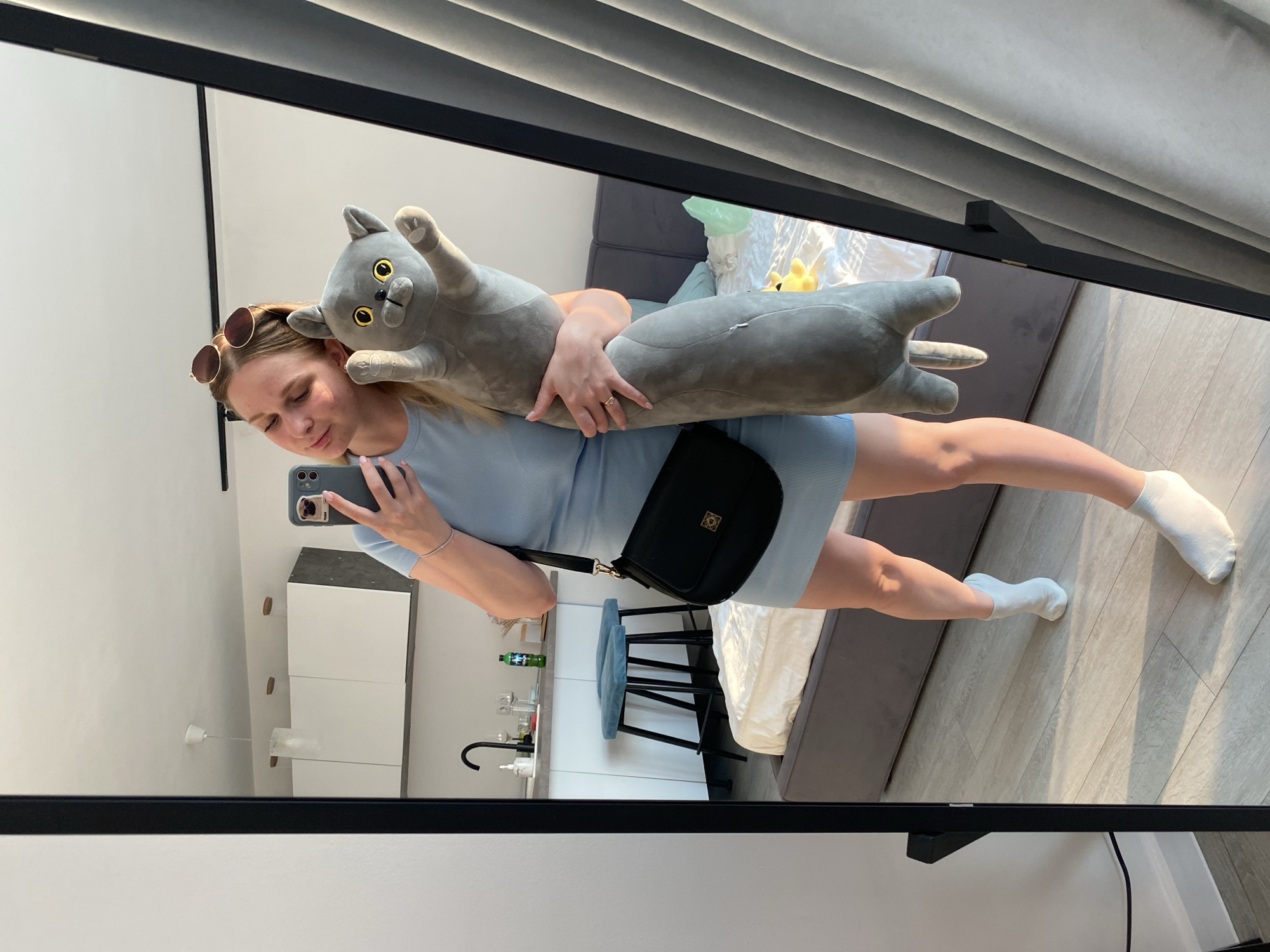 A young woman is captured in a mirror selfie, posing with a large gray stuffed cat. She is wearing a light blue dress and white socks, with sunglasses perched on her head. A black crossbody bag with a gold emblem hangs from her shoulder. In the reflection, a modern apartment interior is visible, featuring white cabinets, a kitchen counter with a sink and faucet, and two blue bar stools. The room is bright, suggesting it's daytime. The woman's expression is a gentle smile, indicating a relaxed and playful mood. The overall scene conveys a sense of domestic comfort and personal style.