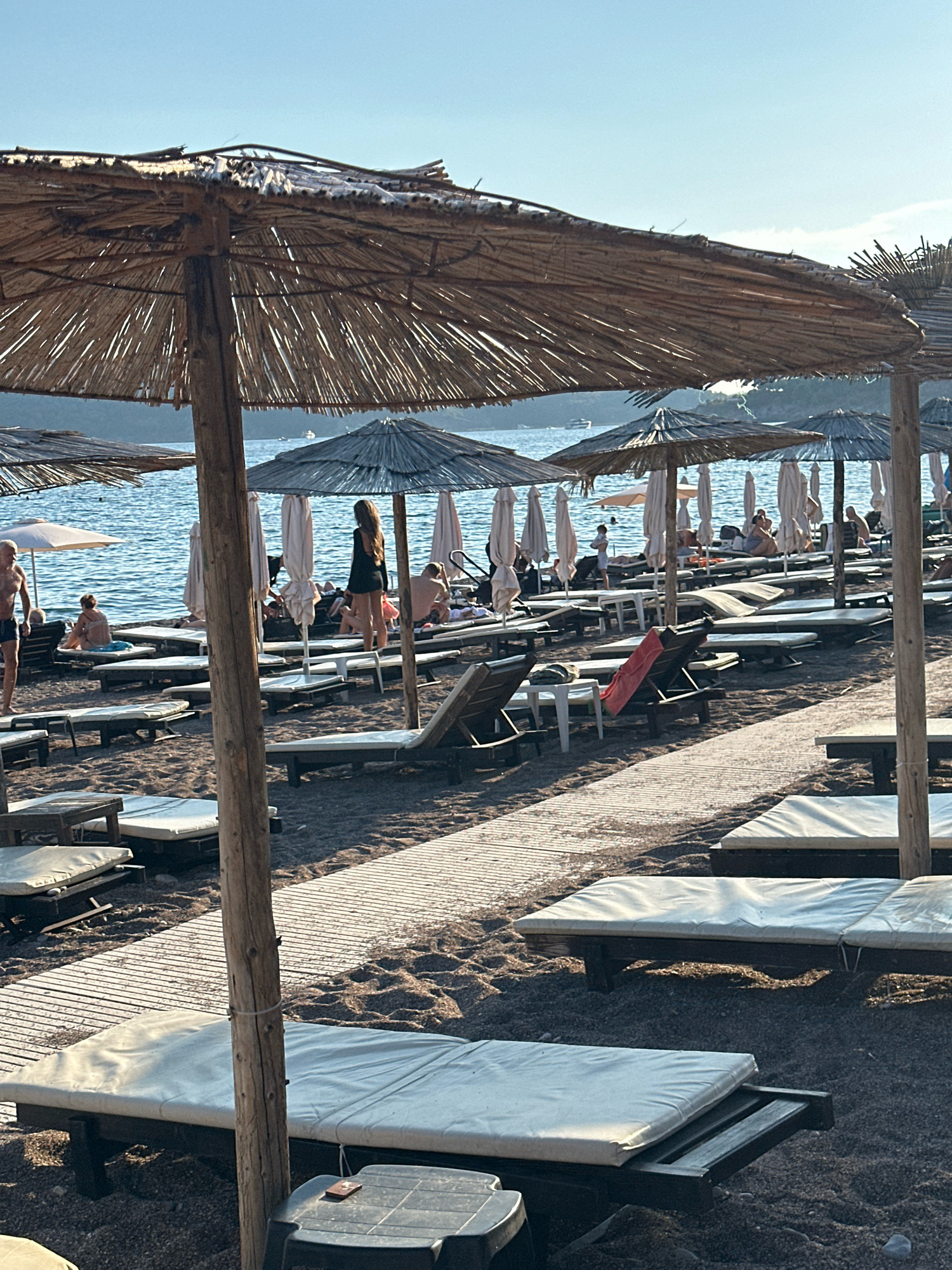 The image depicts a beach scene in Boreti, Montenegro, characterized by numerous straw umbrellas and lounge chairs on a pebble and sand beach. The ocean is visible in the background, with a clear blue sky above. Several people are scattered across the beach, some seated on lounge chairs, others standing or walking. One woman in a dark dress is standing near a straw umbrella, facing away from the camera. Other individuals are partially visible, engaged in typical beach activities such as sunbathing or relaxing. The arrangement of umbrellas and chairs suggests a resort or public beach setting. The lighting indicates it is either early morning or late afternoon, with the sun casting long shadows.