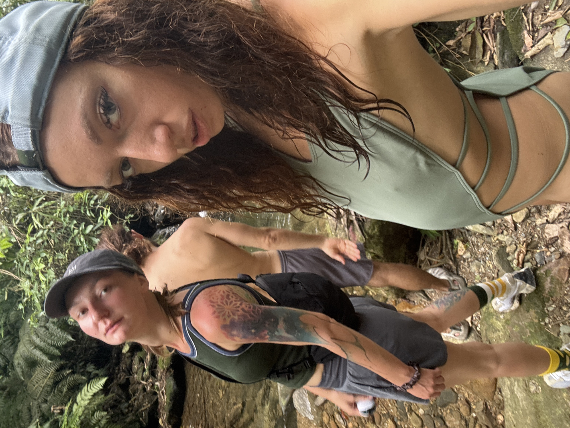 The image shows three people in an outdoor setting, seemingly a natural area. Two women are visible in the foreground. One woman is wearing a light blue baseball cap and a pale green swimsuit-style top with multiple straps on the side. The other woman has a dark baseball cap, a dark green tank top, and a large tattoo on her left arm. She is wearing gray shorts. Behind them is a shirtless man with gray shorts. They appear to be standing near a stream or small waterfall, given the rocks and wet ground. Lush green vegetation and ferns are visible in the background. The photograph is possibly taken in Suối Giàng, Vietnam.