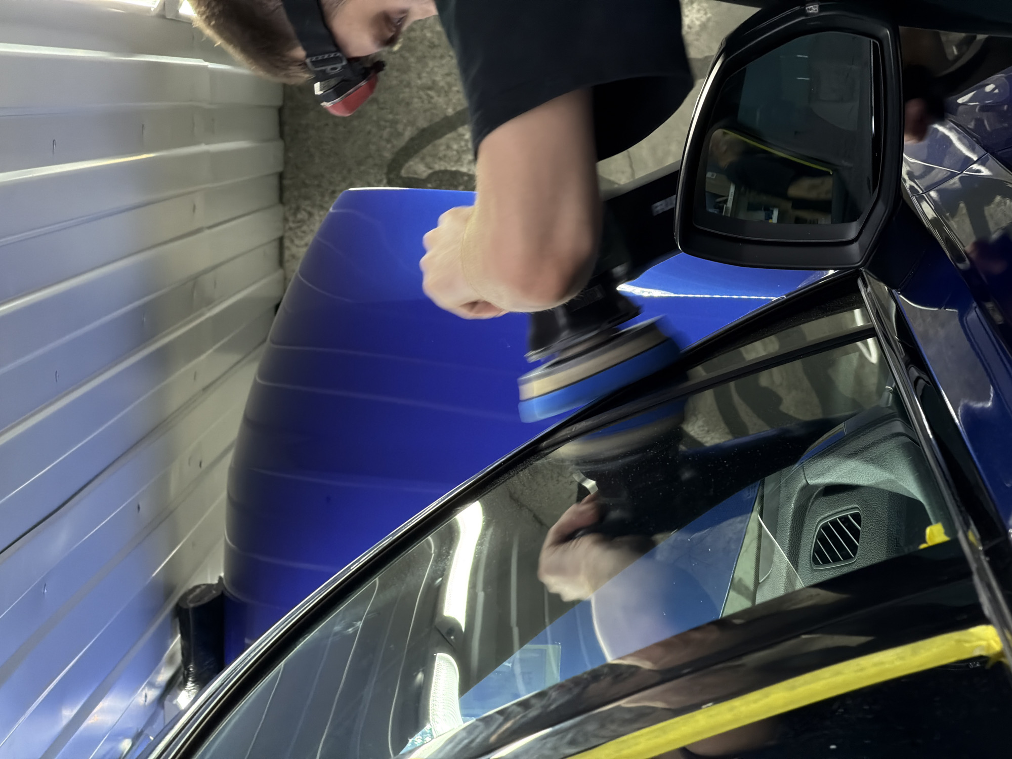 A person is polishing a blue car in what appears to be a garage or detailing shop. The car's side mirror is visible, along with the partially visible interior through the window. Yellow tape is used to protect the edges of the car's panels. The setting is likely indoors, given the presence of walls and artificial lighting. The image offers no specific clues that would locate it in Krasnoyarsk, Russia.