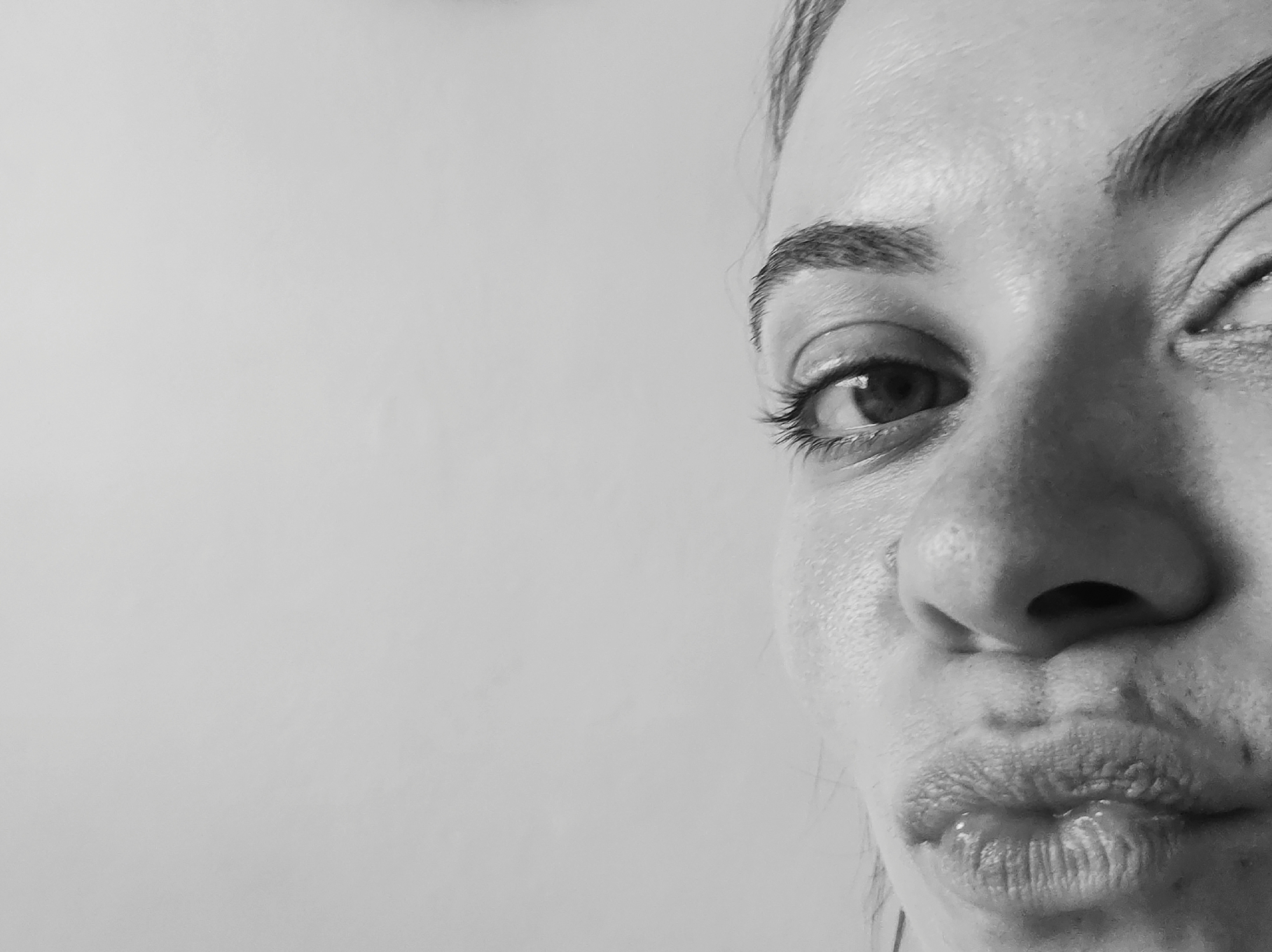 A black and white, tightly cropped portrait features the right side of a person's face, likely female. The subject is captured mid-expression, with her right eye wide open and gazing towards the upper right of the frame. Her lips are noticeably pursed forward, forming a playful or exaggerated "kissing" expression. The left side of her face is partially visible, with her left eye appearing squinted. The background is a plain, light-toned wall, out of focus, suggesting an indoor setting in Chaikovsky, Russia. Notable details include the distinct eyelashes and eyebrow, the texture of the skin, and a slight glisten on the lower lip. The image conveys a sense of playfulness or humor. No text is visible, and the time of day is not discernible.