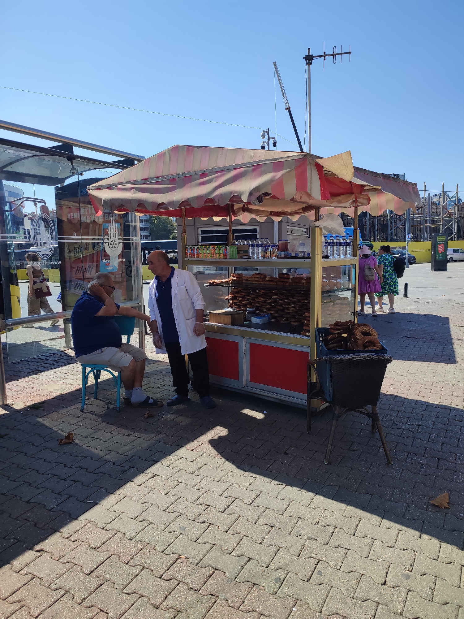 This is a photograph taken outdoors in Kadıköy, Turkey, on a sunny day. In the foreground, a street vendor is selling simit (a Turkish bread). The vendor, a man wearing a white coat, is standing next to his stall, which displays numerous simit on shelves and in a basket. A customer, an elderly man wearing a blue shirt and shorts, is seated on a light blue stool and appears to be engaged in conversation with the vendor. Behind them, a bus stop structure with reflective glass panels is visible. In the background, several other people are walking, and construction scaffolding suggests ongoing building activity. The ground is paved with interlocking bricks, and shadows are cast by the stall and the people. The sky is clear and blue.