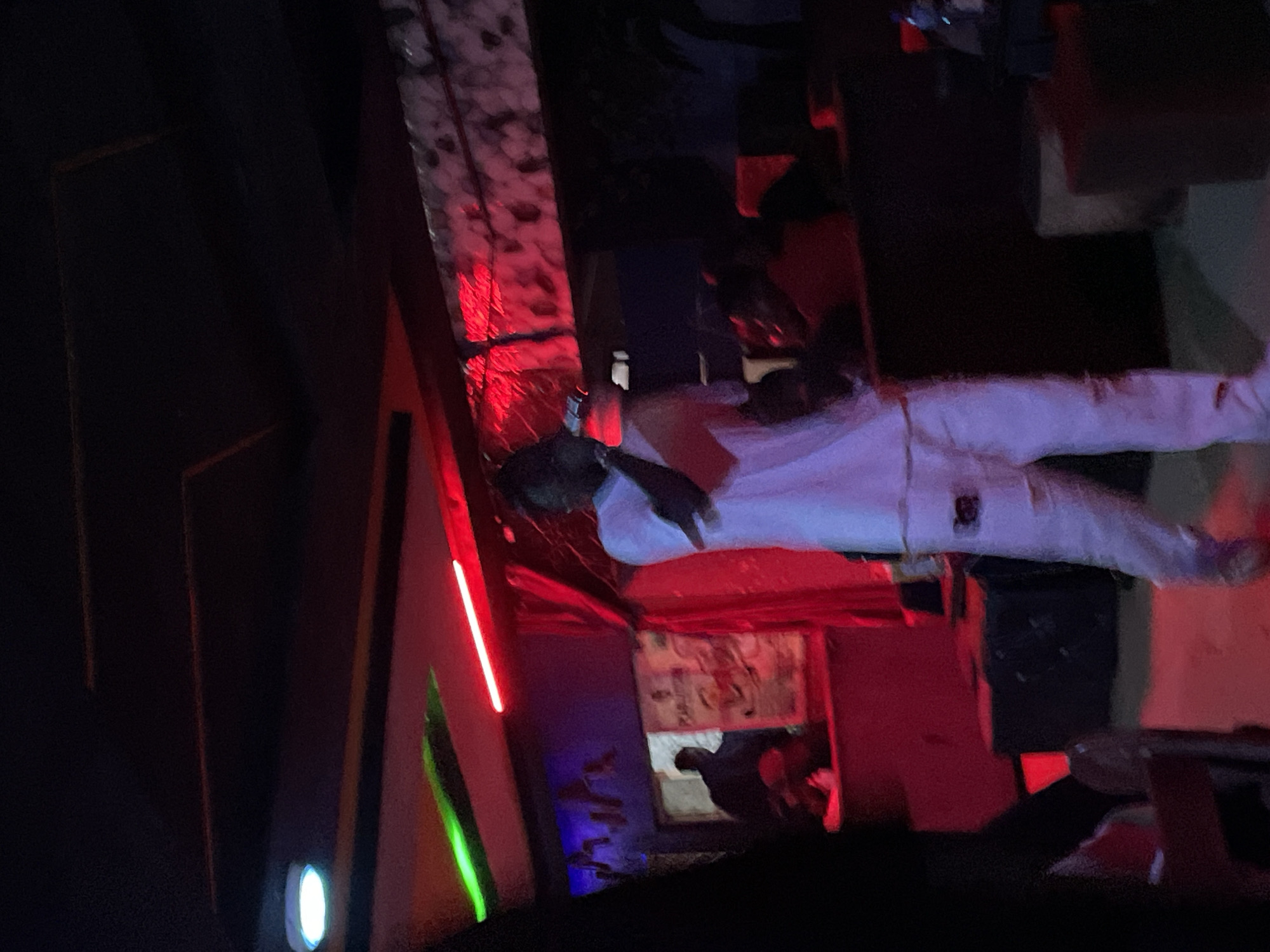 The image depicts a dimly lit indoor venue, possibly a club or performance space in Durumi, Nigeria. A person in the foreground is wearing a white t-shirt and white pants, holding a microphone and appearing to perform or address a crowd. Red and green neon lights illuminate the scene from above, casting a vibrant glow. In the background, several individuals are visible, some in silhouette, and there are various objects that suggest a social gathering, such as tables, seating, and what appears to be a bar area. The overall atmosphere is energetic and celebratory, with the lighting and subject matter indicative of a live entertainment event.