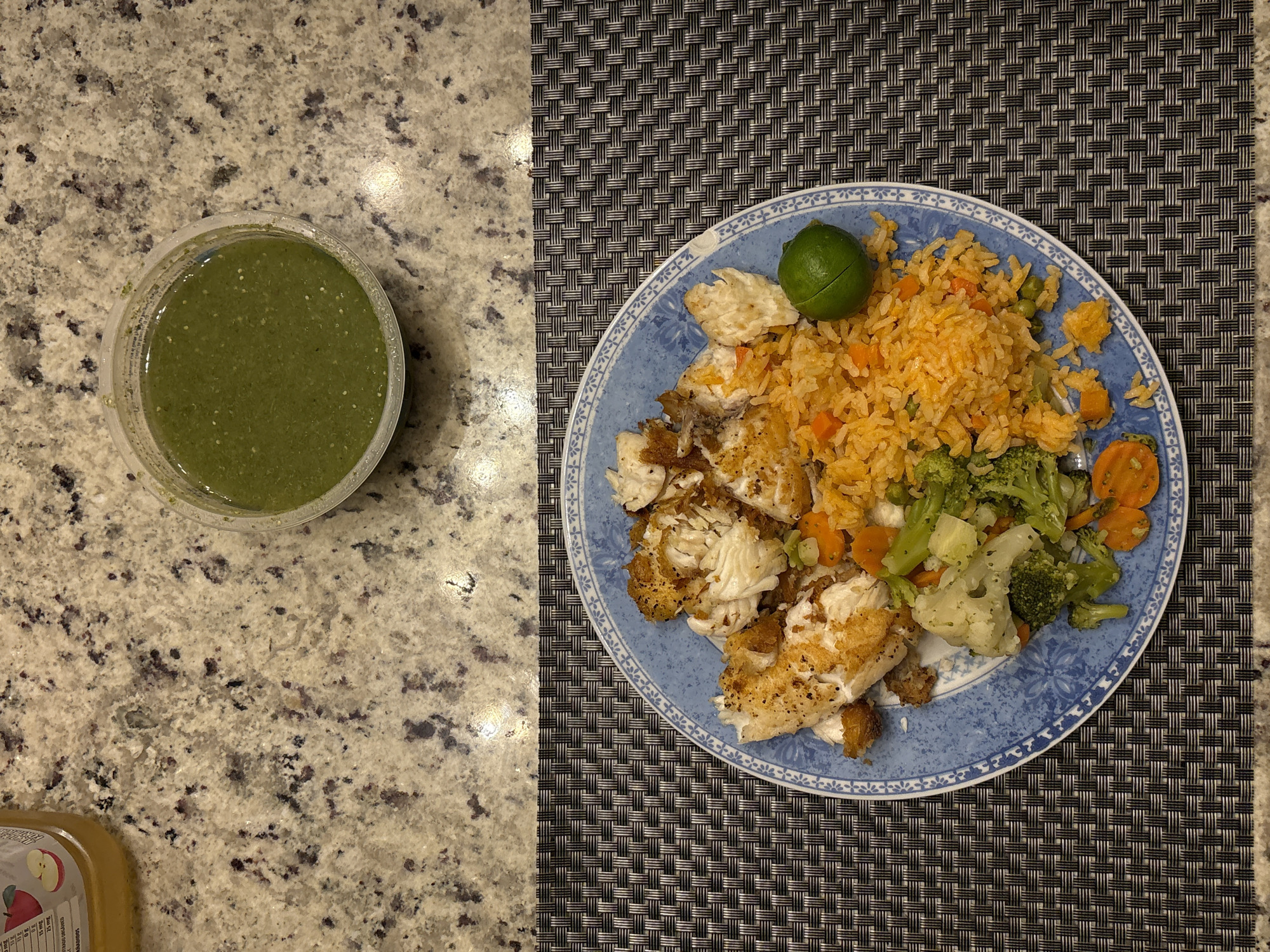 The image shows a meal consisting of fried fish, rice, vegetables (broccoli, cauliflower, carrots), and a lime wedge on a blue patterned plate. A container of green salsa sits next to the plate. The plate rests on a woven black and gray placemat. The salsa container is on a speckled beige countertop. Part of an apple juice container is visible in the lower left corner. There is no contextual information in the image to connect it to San Bernardino Tlaxcalancingo, Mexico.