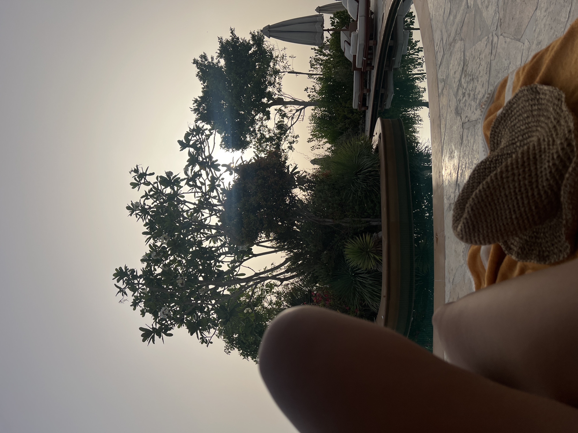 The image depicts a person relaxing by a pool, likely in a resort or private residence. The foreground shows the person's leg and a straw hat resting on a yellow towel. In the background, there is a pool of water reflecting trees, foliage, and beach umbrellas. The sky is bright and hazy, suggesting either dawn or dusk. The scene is most likely set in a luxurious outdoor location, with details suggesting a resort. While I cannot definitively confirm the location is Dubai, United Arab Emirates, the presence of a resort-style pool, the type of vegetation (such as palm trees), and the quality of light would support the assertion that it could be the location.