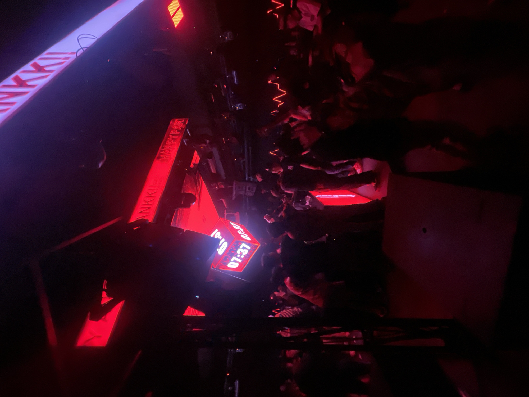 The image depicts a dimly lit interior, likely a nightclub or event space, bathed in predominantly red and purple neon lighting. In the foreground, a crowd of people, mostly silhouetted due to the low light conditions, are gathered. Several individuals are in motion, suggesting dancing or active socializing. Overhead, a large, cubic digital display shows the time "07:37" on multiple faces. Above this, a bright white and purple illuminated sign with red lettering reads "ANKK!". Further signs with red illumination are visible, with one displaying "HAPPY PART". The environment appears to be within a building, possibly a commercial establishment in North Jakarta, Indonesia, given the context provided. The overall atmosphere is energetic and characteristic of a nightlife setting.