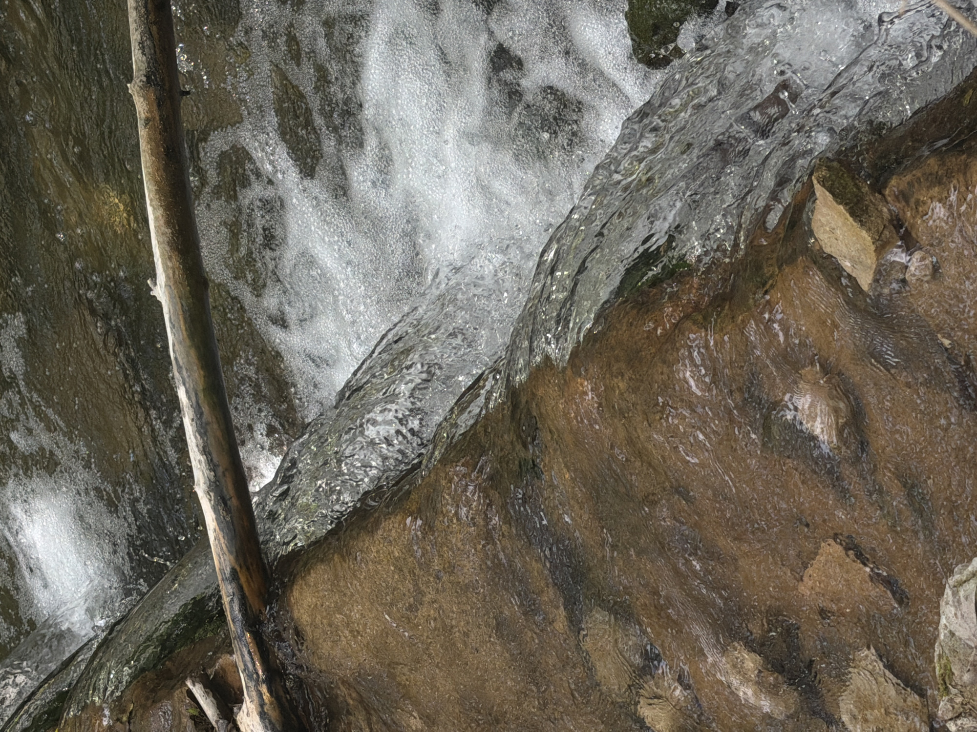 This close-up image captures a natural scene of water flowing over rocks and around a piece of wood, likely part of a stream or small cascade in Sant Martí Sarroca, Spain.
The primary subject is the dynamic movement of water. In the upper left and central areas, the water is turbulent and highly aerated, appearing white and foamy with numerous bubbles as it rushes downwards. Below this, the water flows more smoothly over the contours of the rocks, revealing its clarity and the textures beneath. The surface of this clearer water shimmers with reflections, indicating light interacting with its movement.
A prominent feature is a thick, dark brown branch or log, positioned vertically on the left side of the frame. It appears smooth and wet, partially submerged in the flowing water. To the right and bottom, rugged rock formations dominate, showing a variety of earthy tones from light brown and tan to darker, rich reddish-browns, with some darker patches that could be dampness or moss. The rocks exhibit visible stratification and wear from the water's flow.
The activity depicted is the continuous flow of water through a natural, rocky environment. The lighting suggests it is daytime, with diffused light possibly due to an overcast sky or a shaded location, as there are no harsh shadows. The scene evokes a sense of natural tranquility and the persistent power of water shaping the landscape. No text is visible in the image.