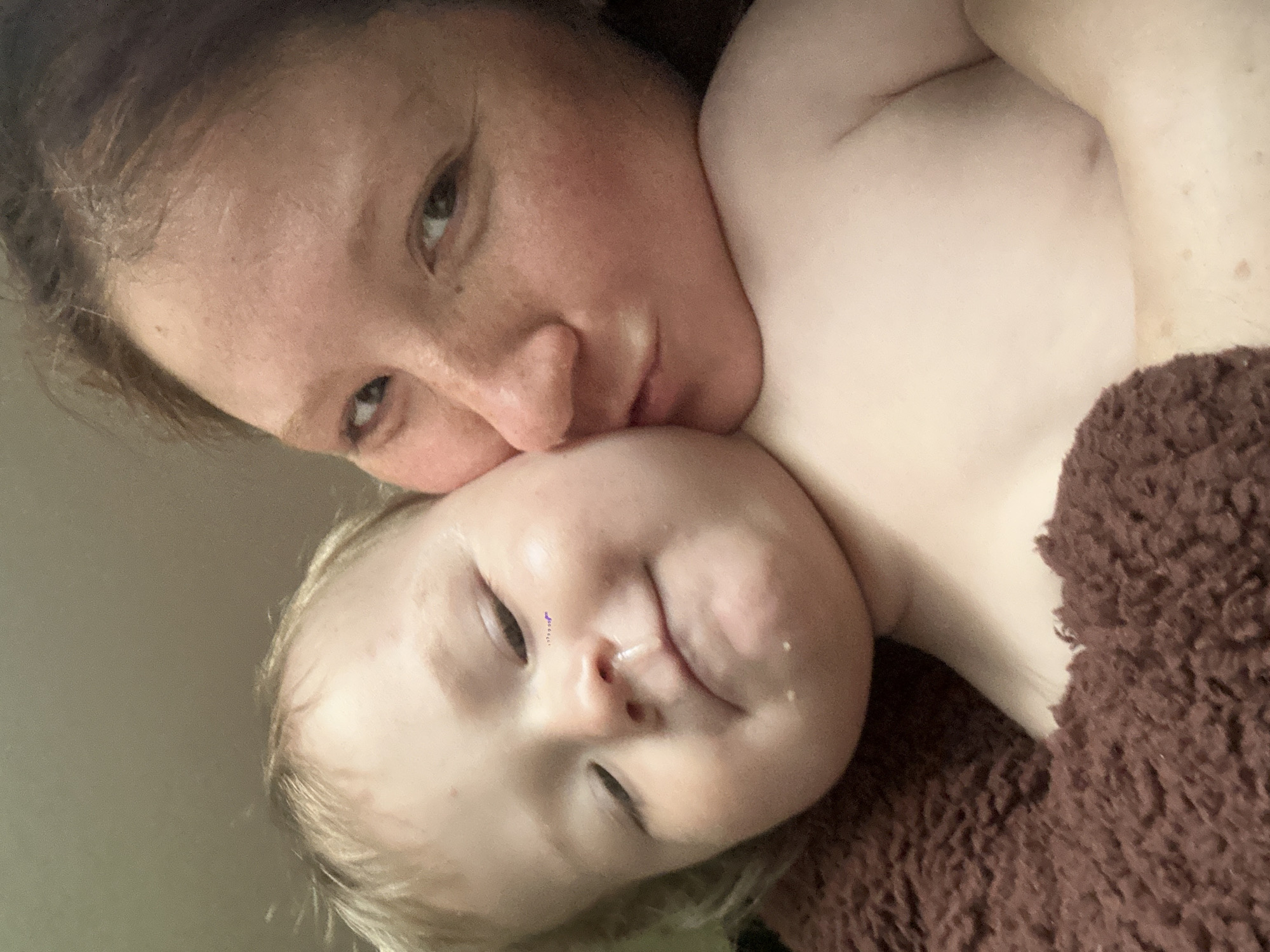 A woman is closely nuzzling a young child. The child is lying on a brown, textured blanket or surface. The woman's face is close to the child's cheek, with her eyes visible and a focused expression. The child's eyes appear closed or squinting, with a small amount of liquid visible on their face. No elements suggest the location is Nizhny Novgorod, Russia.