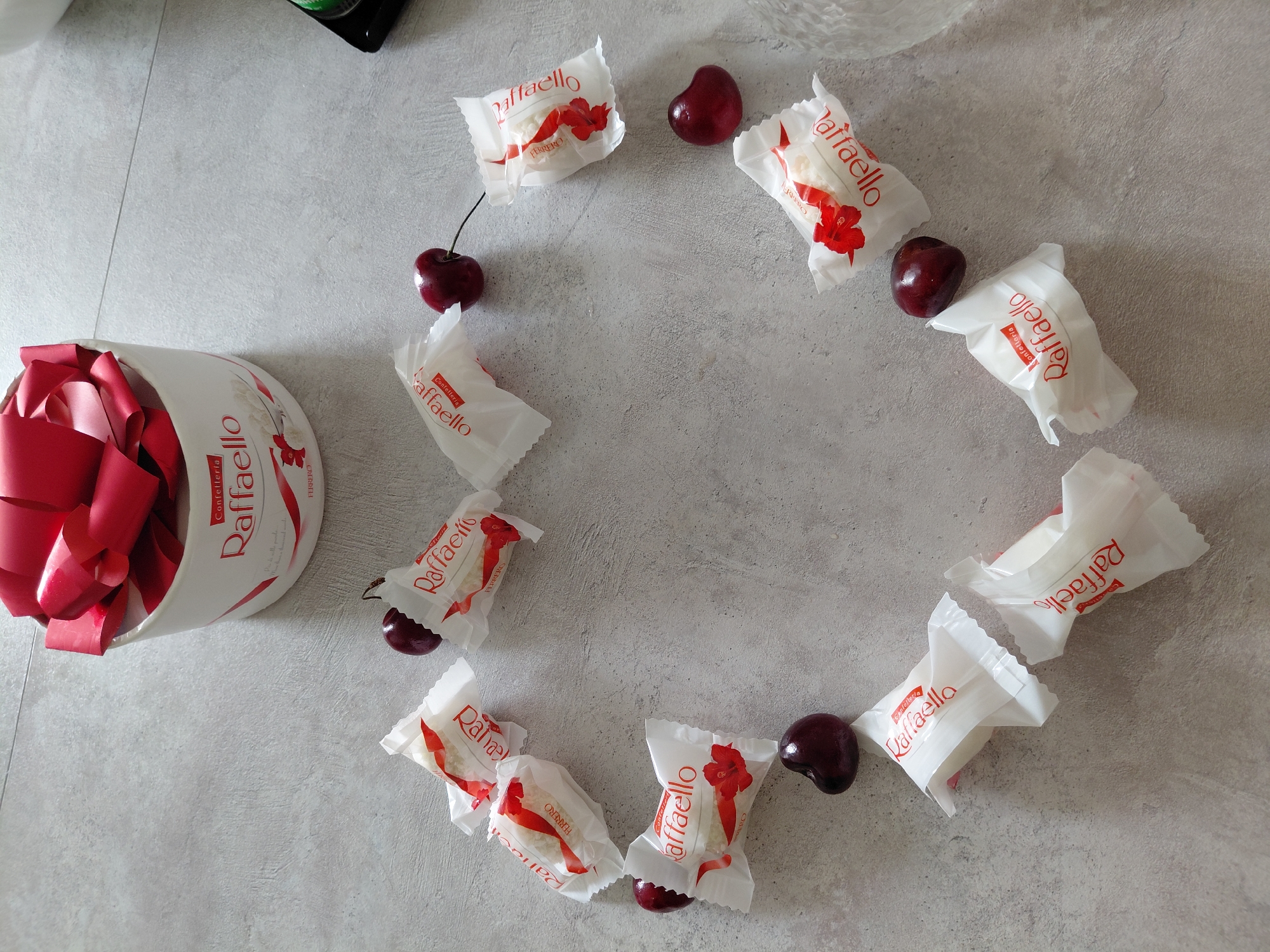 The image shows a collection of RaffaELLO chocolates and cherries arranged in a heart shape on a grey, textured surface. At the top of the image, a white cylindrical box with a red ribbon is visible, also containing RaffaELLO chocolates. The overall arrangement suggests a festive or romantic theme. The scene is indoors, with neutral lighting. The location in Novosibirsk, Russia, is not determinable from the image itself.