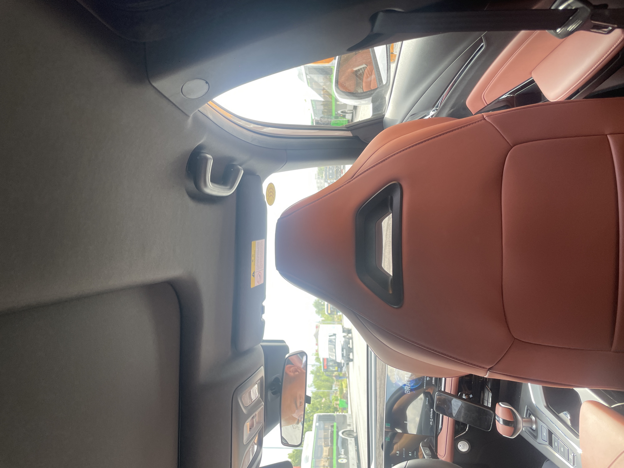 The image shows the interior of a car, looking forward from the back seat. The car has brown leather seats and a sunroof. In the foreground, the headrest of the driver's seat is visible. The dashboard and steering wheel are also visible, as is a smartphone mounted on the dashboard. Outside the car, traffic can be seen, including buses and a white truck. The scene appears to be a city street, possibly in Almaty, Kazakhstan, given the presence of traffic and buses. The overall lighting suggests it is daytime.