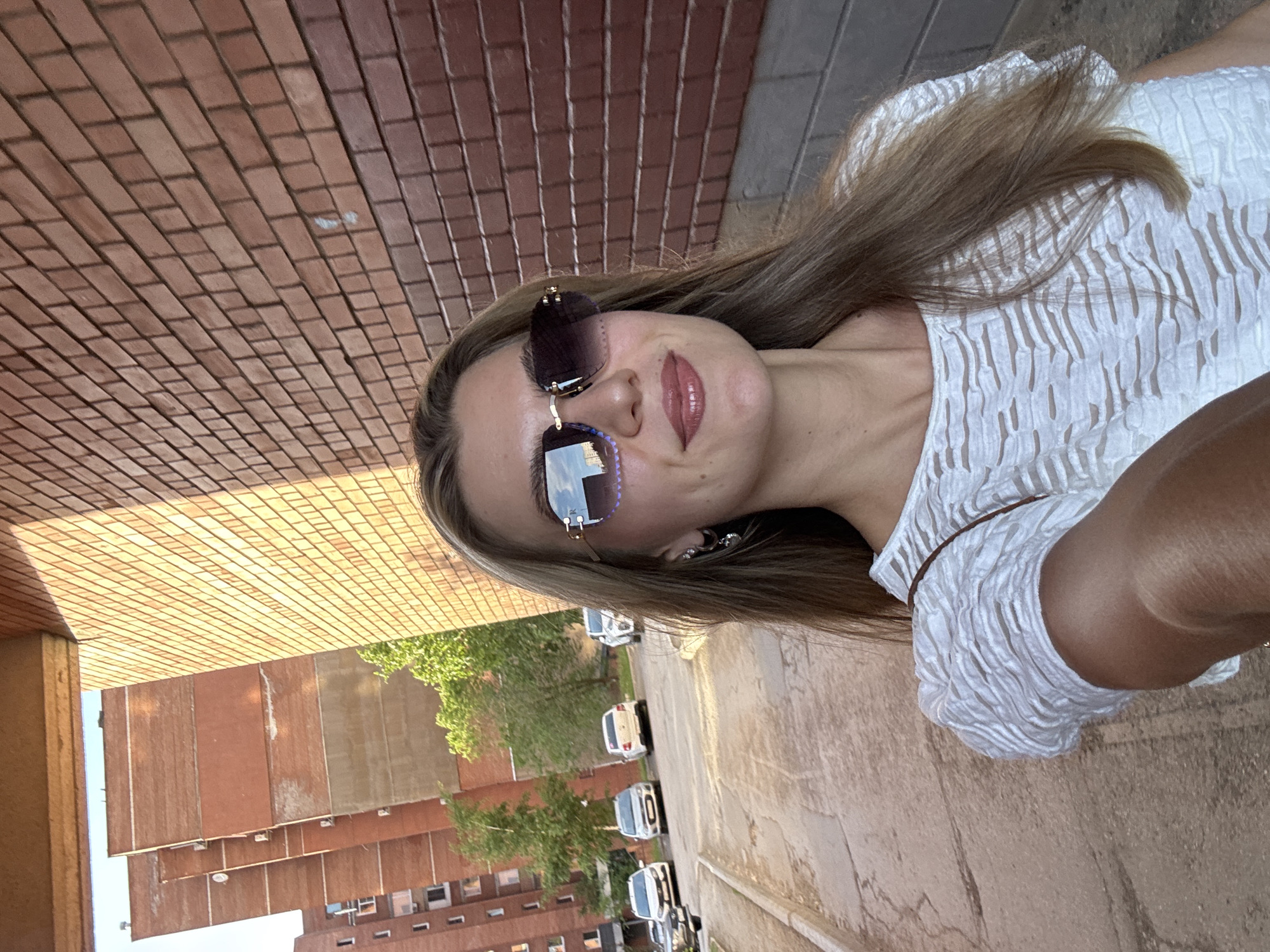 A young woman with long blonde hair is standing outside in a city, wearing sunglasses and a white textured top. She is facing the camera, holding it in her extended right arm. Behind her is a brick wall, a street lined with parked cars, trees, and a tall brick building with multiple windows. The city appears to be in Unknown, Unknown.