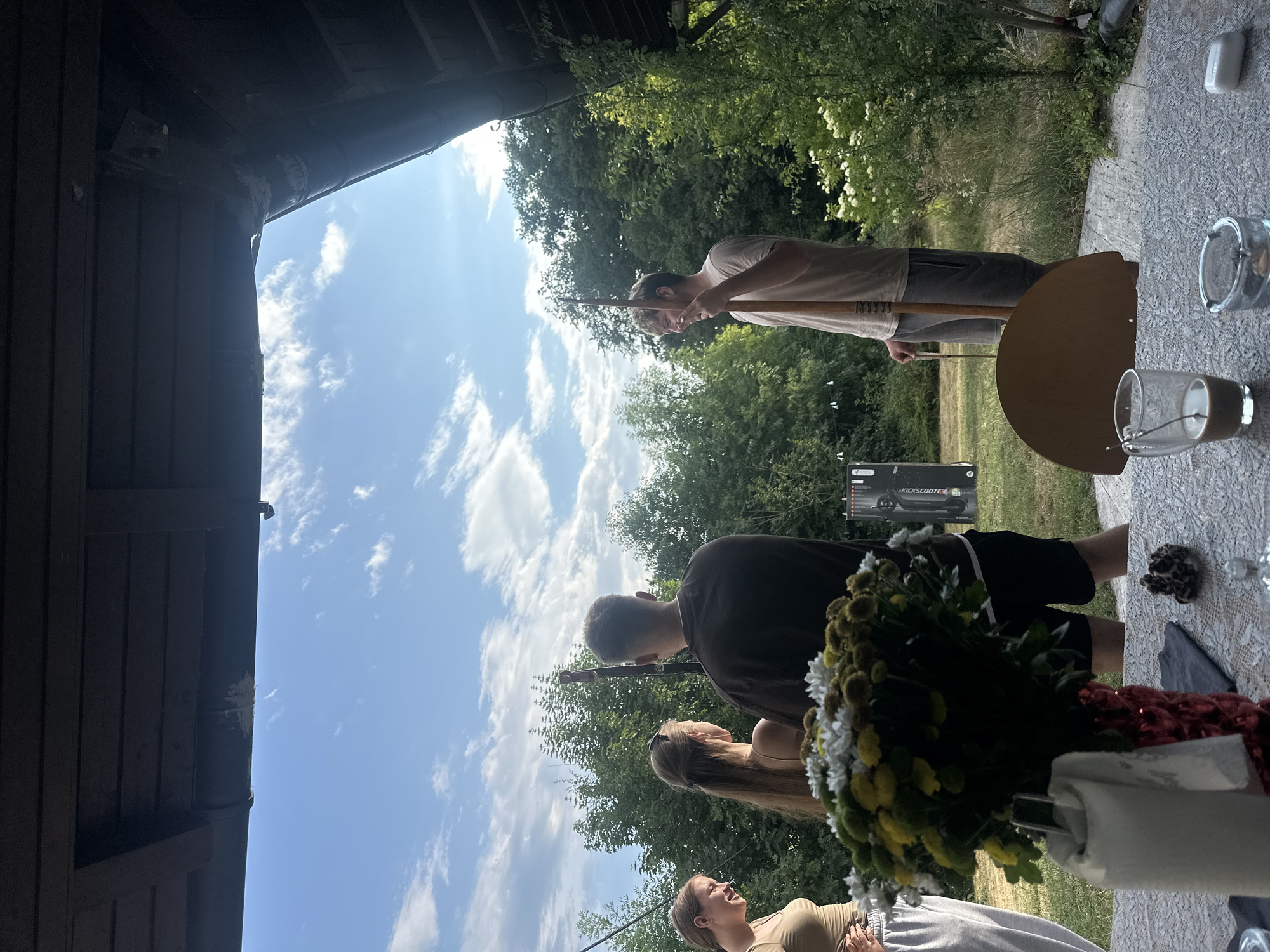 The image depicts a gathering of four individuals outdoors, possibly in a backyard or patio area. The scene is viewed from beneath a wooden structure, likely a porch roof. In the foreground, a table is set with a lace tablecloth, a vase of flowers, a glass of dark liquid, an ashtray, and a bar of soap.
There are three women and one man visible. On the far left, a woman wearing a tan top and white pants is partially visible, smiling. Next to her, another woman with long brown hair, wearing a tan tank top, faces away from the camera. A man with short brown hair, wearing a dark sweatshirt and black shorts, stands between them, also facing away. On the right side of the image, a man wearing a light-colored t-shirt and grey shorts is holding a long wooden staff and looking to his right with a smile.
In the background, lush green trees and bushes form a natural backdrop under a partly cloudy blue sky. There is also a black, flat object leaning against a tree or structure in the middle-ground, which appears to be a poster or a large piece of equipment. The overall atmosphere suggests a casual, social event in a suburban or rural setting. The presence of the staff suggests a potential activity related to archery or a similar hobby. The time of day appears to be late afternoon or early evening, judging by the lighting. There is no explicit indication of the city of Günzburg, Germany, within the image itself.