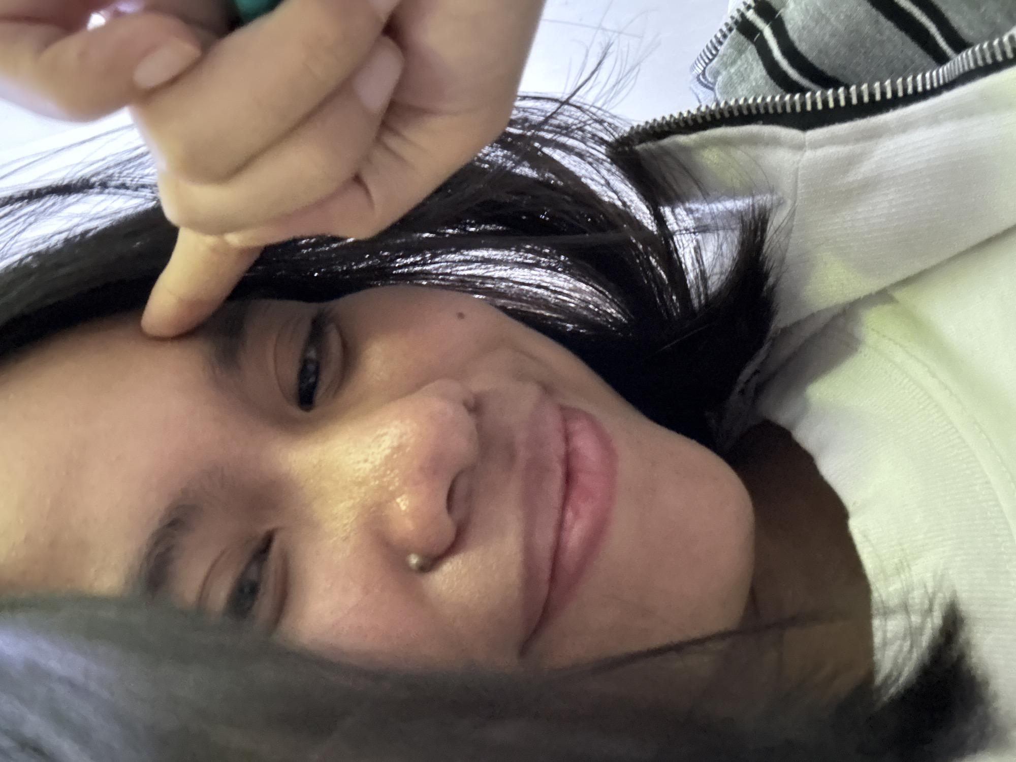 A close-up selfie shows a young woman with dark hair, a nose piercing, and fair skin. She is wearing a white t-shirt and a grey and black striped jacket. She is pointing to her forehead with her right index finger. The background is blurred but appears to be an indoor setting with white walls and possibly a window. There is no visible indication of the location within East Jakarta, Indonesia.