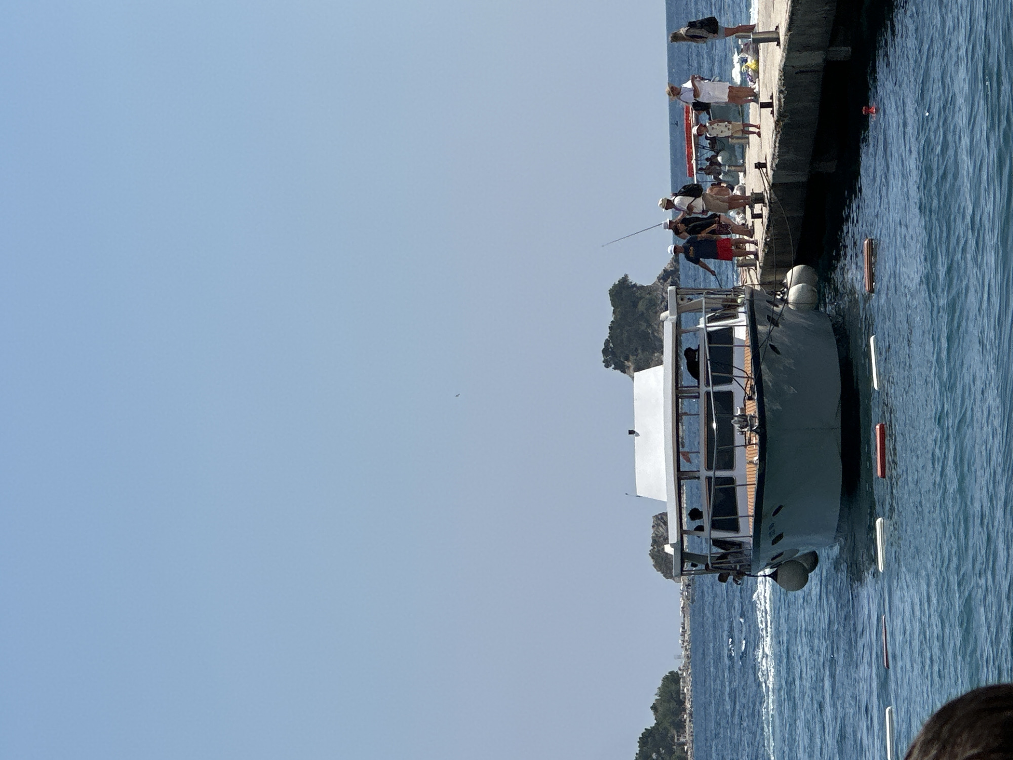 The image depicts a sunny day at a pier in Budva, Montenegro, with the clear blue sky dominating the upper portion. Several boats are docked or visible in the water. In the foreground, the bow of a white boat is prominent, with two large white fenders attached to its side. A concrete pier extends into the water on the right side of the frame, where a group of people are gathered.
On the pier, there are approximately eight individuals. Two men are standing near the edge, one wearing a dark blue t-shirt and red shorts, holding what appears to be a fishing rod. Another man, wearing a white shirt and shorts, is also near the water. A woman in a white dress stands further down the pier, facing the water, and another woman in a dark jacket and skirt is at the end of the pier. Several other people, including a child, are also visible on the pier, some appearing to be interacting or waiting.
In the background, the coastline of Budva is visible, characterized by lush green trees and some buildings. A distinctive rocky island formation rises from the sea. The water is a vibrant blue, with gentle waves and some floating markers. The overall scene suggests a tourist or recreational area.