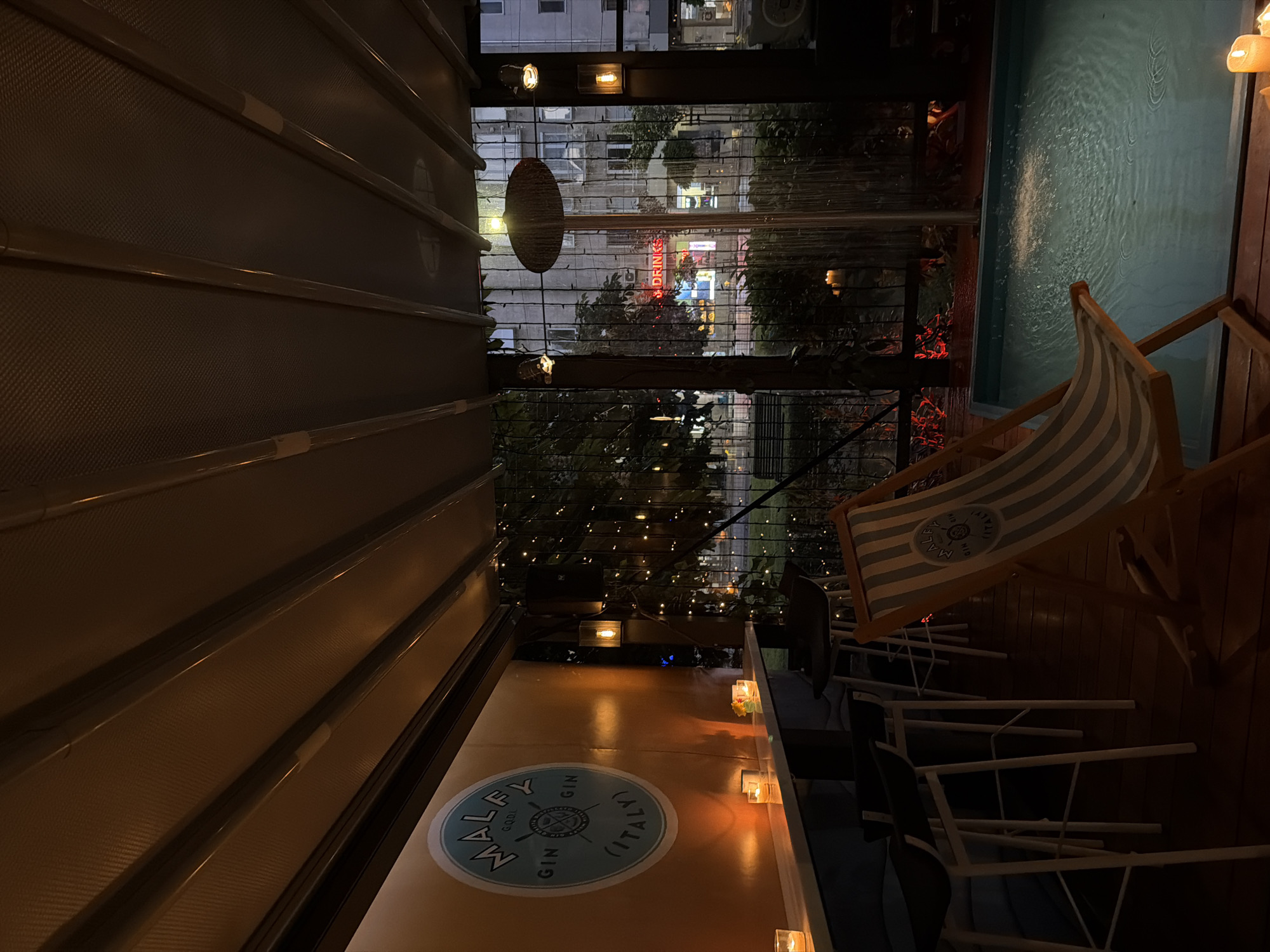 The image shows a dimly lit outdoor space. A partially visible, illuminated water feature is present on the right. A striped beach chair sits nearby, with a logo reading "MALFY Gin, G.D.I. (Italy)" displayed. Several chairs and tables are stacked up beside it. In the center of the scene, a view to an outdoor space with trees and the word "drinks" written with neon lights, which provides a backdrop, is visible through what appears to be a lattice screen. A large logo displaying "MALFY Gin G.D.I. (Italy)" is emblazoned on the surface. The image suggests an outdoor bar or lounge setting. The scene location is Sofia, Bulgaria.