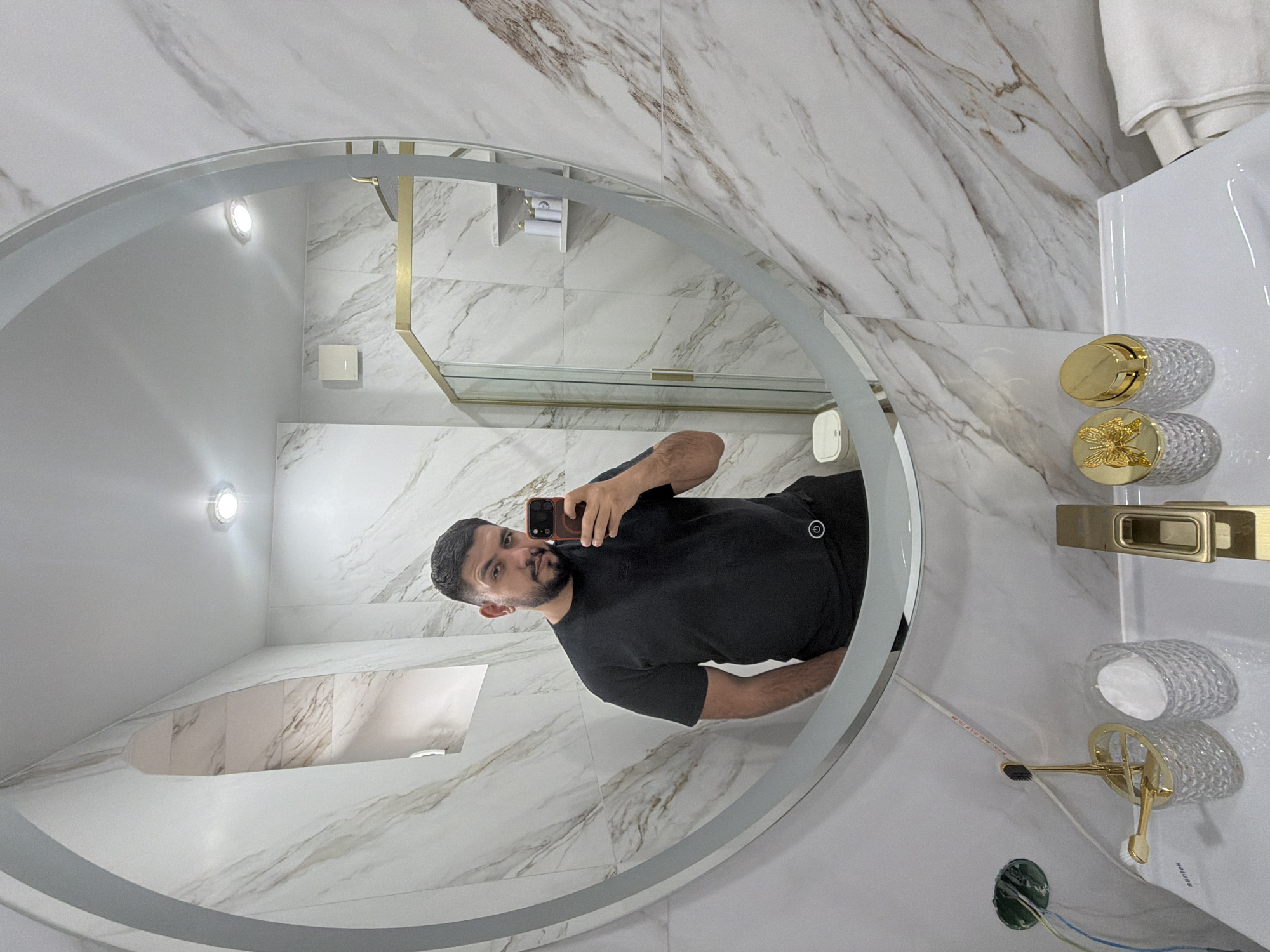 A man is reflected in a round mirror in what appears to be a bathroom. The man is holding a smartphone up to take a selfie. He has dark hair, a beard, and is wearing a black t-shirt.
The bathroom is decorated in white and marble. A gold-trimmed shower stall can be seen in the background. A sink is visible on the right, with ornate gold and glass fixtures, including a toothbrush holder. There is an open electrical outlet on the sink counter.
The reflection in the mirror suggests that this space could be located in Krasnoyarsk, Russia, assuming that the person taking the photo is a resident of that city. This relies on the assumption that people typically take selfies in their own bathrooms or residences. However, this is speculation, and the photo does not provide conclusive evidence of the location.