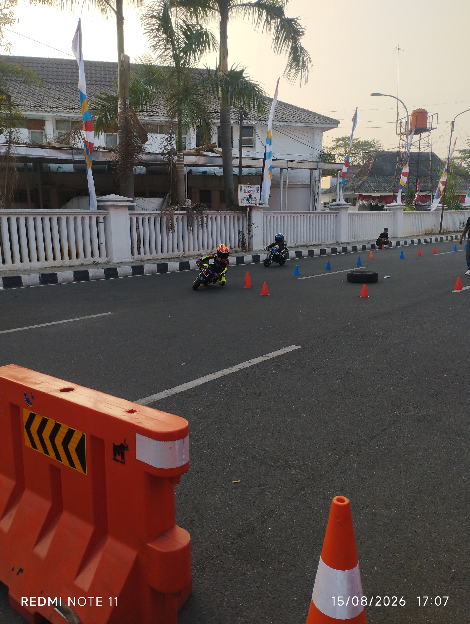 The image captures a motor racing event, likely a children's or amateur race, taking place on a street in Karawang, Indonesia. The foreground features an orange traffic barrier with reflective chevron markings and a branding logo. The road is marked with white lines and dotted with orange and blue traffic cones, indicating a course or obstacle. In the mid-ground, two young racers on miniature motorcycles are actively participating in the event. One racer, wearing a red and yellow helmet and a black and yellow racing suit, is leaning into a turn. Another, in a blue helmet and a blue and white racing suit, is further down the course. Spectators are visible along the roadside, including a person crouching near the cones. A white fence with decorative posts separates the racing area from buildings in the background. These buildings appear to be commercial or residential, with a prominent water tower visible in the distance. Several flags, including Indonesian flags and decorative banners, are displayed along the fence. The sky is clear and shows the warm hues of either sunrise or sunset. The timestamp at the bottom right indicates the image was taken on August 15, 2026, at 17:07.