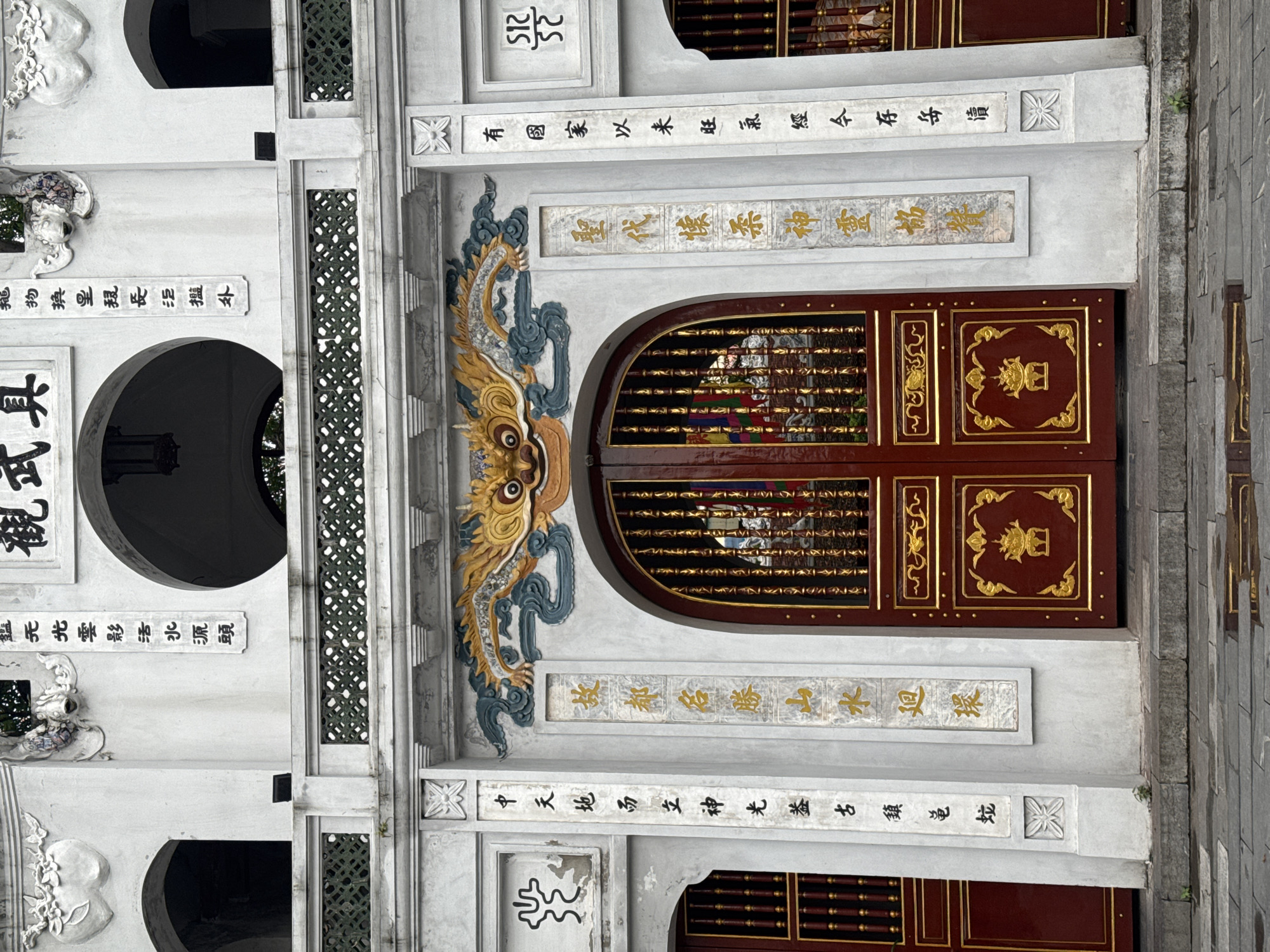 This media file captures a section of an ornate building, likely a temple or a historic structure, in Hà Nội, Vietnam. The building features a white, textured facade with intricate carvings and inscriptions. A prominent element is a large, arched wooden door with a dark red finish and elaborate gold trim. The door is divided into panels, each decorated with golden motifs. Above the door, a striking dragon-like carving in shades of blue, yellow, and gold, with detailed scales and a fierce expression, is mounted on the facade.
Above and around the door, there are horizontal bands of inscribed text in what appears to be traditional East Asian script, some in black ink and others in gold. These inscriptions add to the cultural and historical significance of the structure. Several circular openings, dark within, are visible on the left side of the facade, hinting at interior spaces or architectural features. The building's surfaces are adorned with various decorative elements, including floral carvings and geometric patterns. The ground to the right of the building is paved with grey stone tiles. The lighting suggests it is daytime, with no particular weather conditions immediately apparent. The overall impression is one of historical depth and cultural richness.