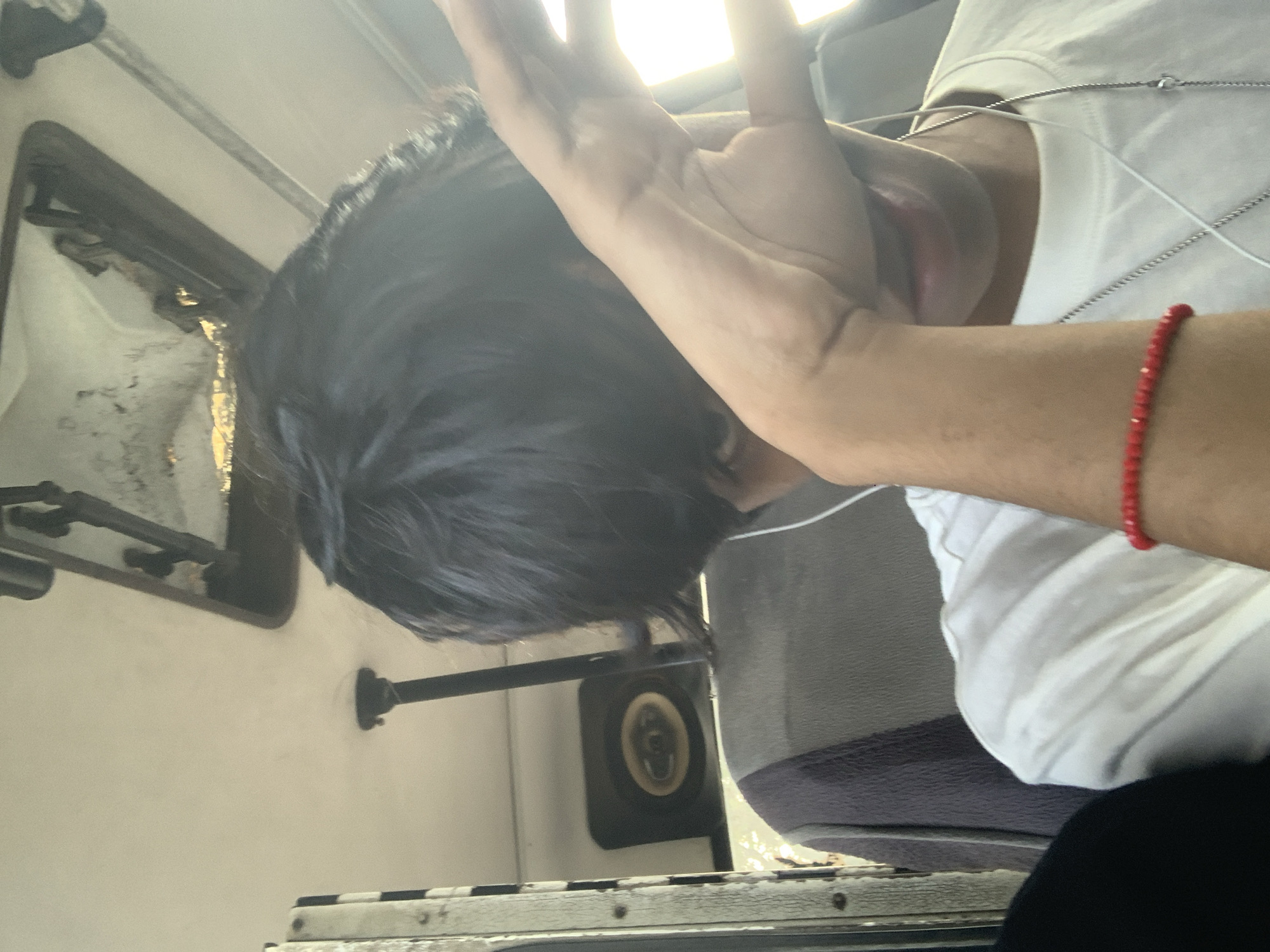 An oblique, vertically oriented photograph captures a person inside what appears to be a moving vehicle, likely a bus or van, during daytime. The individual, seen from a high angle, has dark, short hair and is wearing a white t-shirt. Their right hand is raised, palm facing outwards, partially obscuring their face and shielding from a bright light source coming from the upper right, which creates a strong backlight. Part of their mouth is visible, slightly open.
Notable accessories include a red beaded bracelet on their right wrist and at least one thin silver chain necklace around their neck. White earphone wires are also visible, draped around their neck and chest.
The interior of the vehicle is visible around the person. On the left, parts of a window frame and the vehicle's ceiling or upper paneling are seen, showing some wear and tear. A bright light reflects off surfaces near the window. Towards the bottom center, a circular speaker is mounted on a dark panel. Parts of a seat or cushion, possibly grey or dark fabric, are also visible. The overall setting suggests a functional, possibly older, public or transport vehicle. No visible text is present. Given the context, this scene is located in Porlamar, Venezuela.