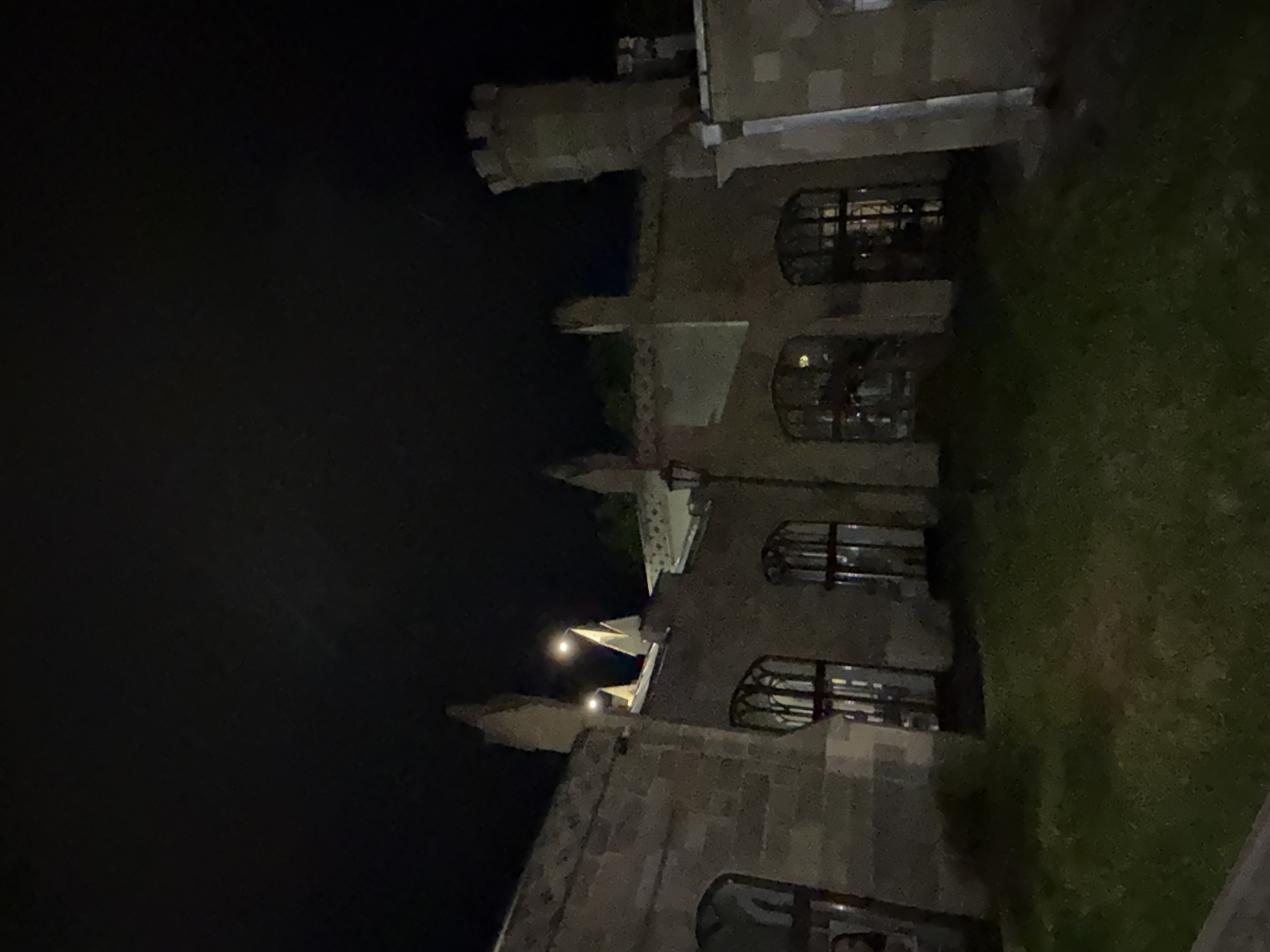 The image shows a stone building with gothic architectural elements, illuminated by artificial lights at night. The building has arched windows with intricate tracery and crenellated turrets. A lamppost stands in front of the building, and a patch of grass is visible in the foreground. The sky is dark, suggesting nighttime. This building is identified as the Narzan Gallery in Kislovodsk, Russia, a well-known architectural landmark.