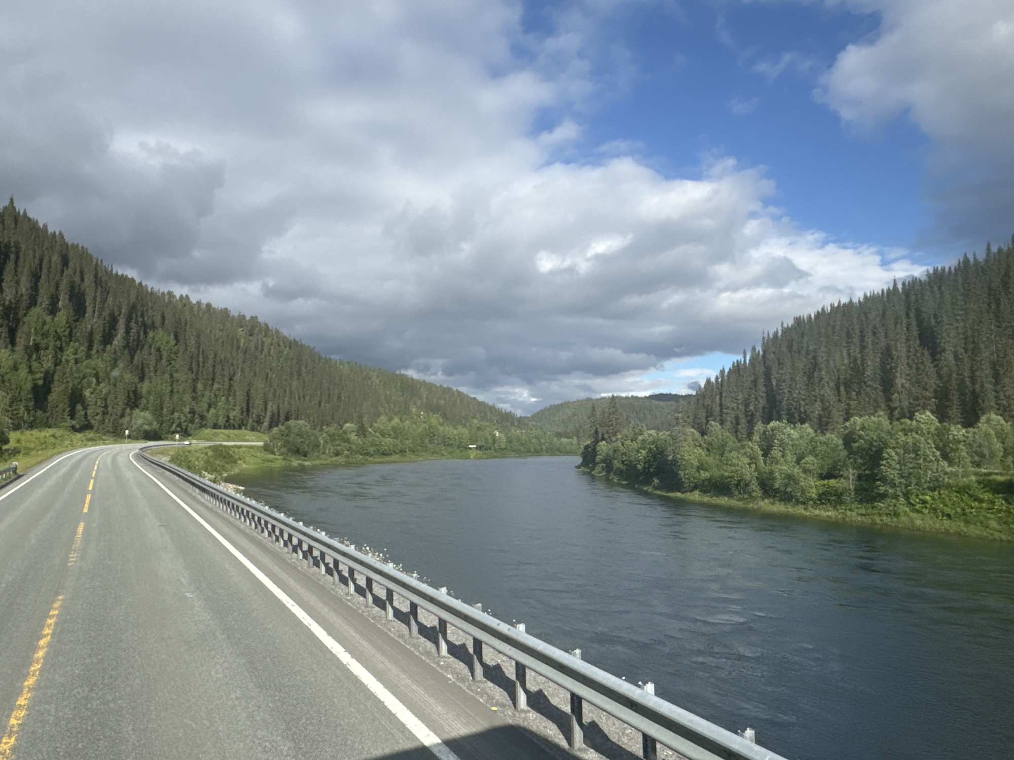 This is a wide shot of a road curving alongside a wide, dark river. The road is asphalt with yellow and white lane markings. A metal guardrail runs along the edge of the road, separating it from the river. The river is flanked by steep, densely forested hills. The trees are a mix of coniferous evergreens and deciduous trees, with the evergreens forming the dominant feature of the landscape. The sky is partly cloudy, with dramatic, dark gray clouds mixed with patches of bright blue sky. Sunlight breaks through in places, illuminating parts of the landscape. The overall impression is one of a remote, natural setting, possibly in a northern climate due to the dense evergreen forests. The time of day appears to be late afternoon or early evening, given the quality of the light and the shadows. There are no people or animals visible in the image. The location is Røssvoll, Norway, suggesting a mountainous, fjord-like landscape. The mood is serene and somewhat dramatic, with the contrast between the natural beauty and the vast, imposing sky. No text is visible in the image.